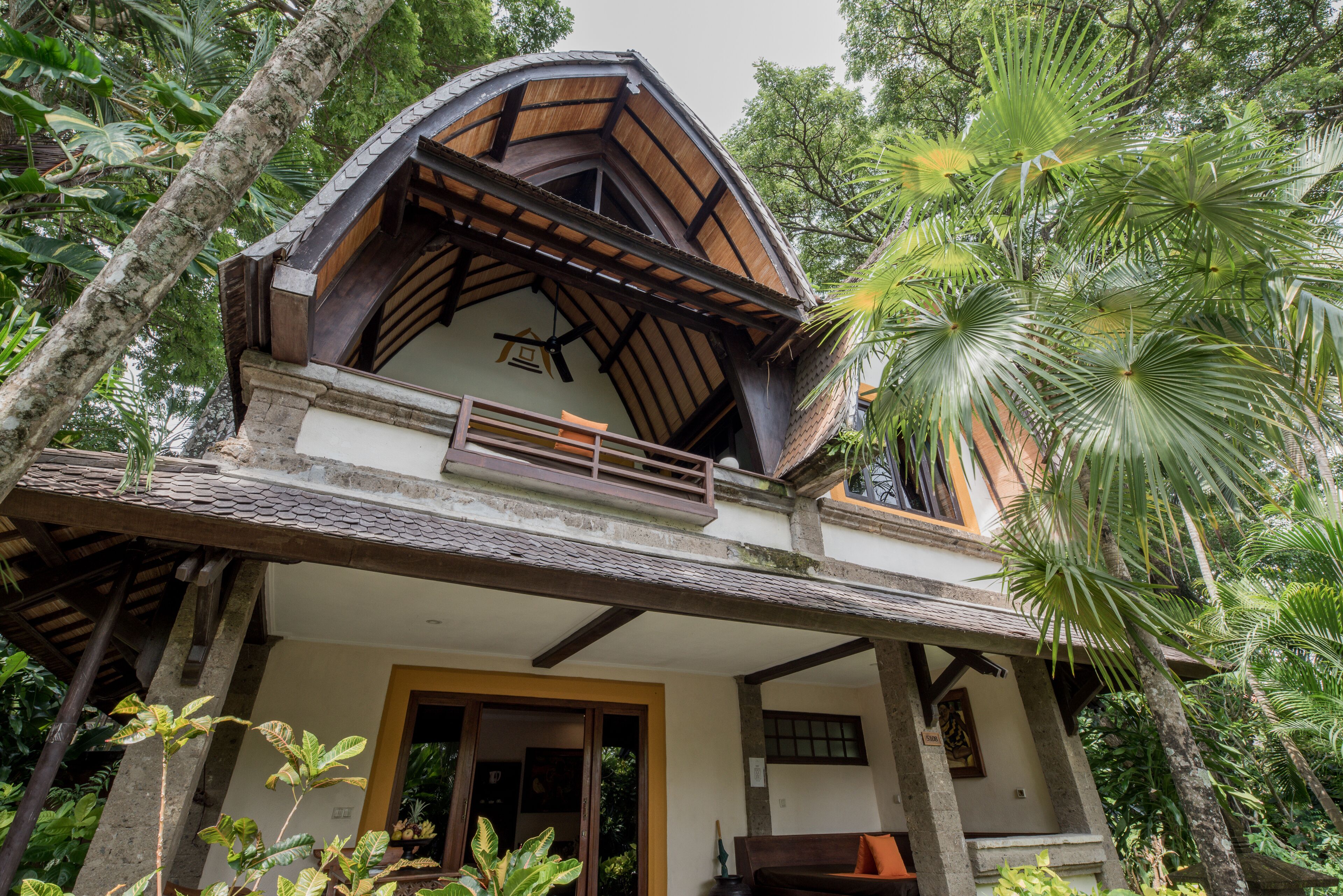 A two-story house with a balcony, surrounded by lush greenery.
