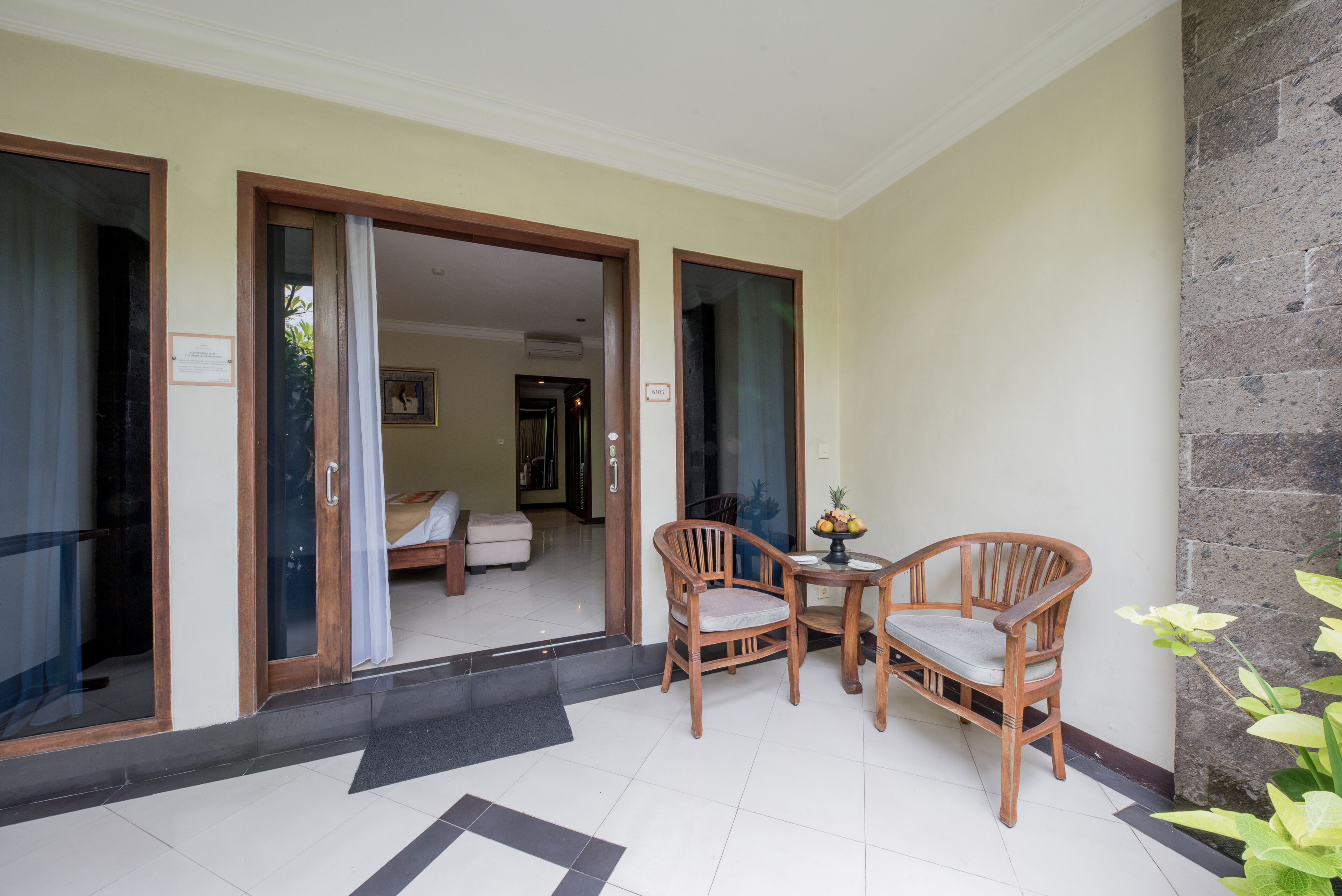 L’entrée d’un hôtel avec un sol carrelé, des chaises en bois, une petite table sur laquelle se trouve un panier de fruits, et on peut apercevoir un lit à l’intérieur.