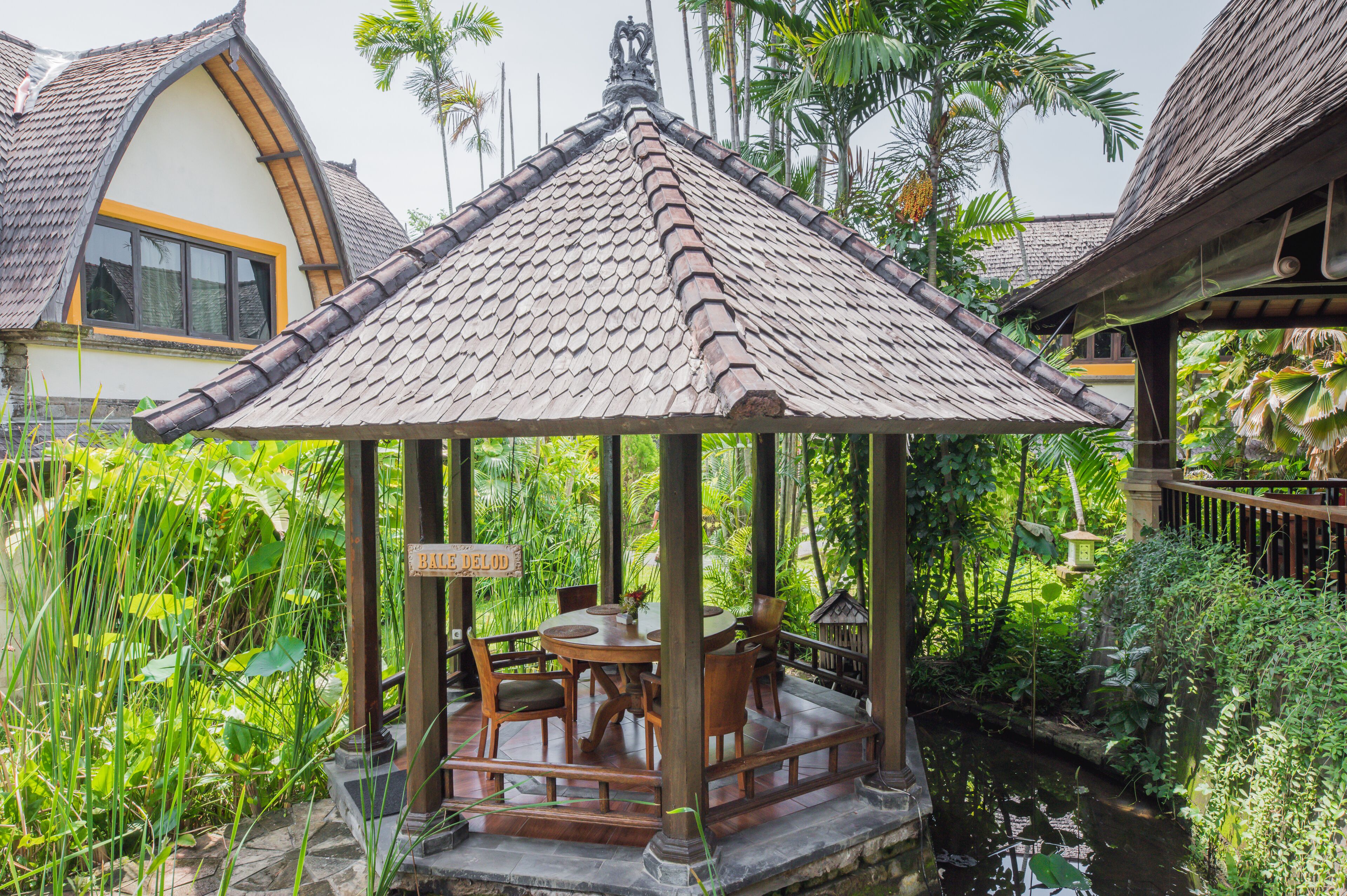 Un belvédère traditionnel en bois, doté d’un toit en tuiles, entouré d’une végétation luxuriante et agrémenté d’un petit élément décoratif aquatique.