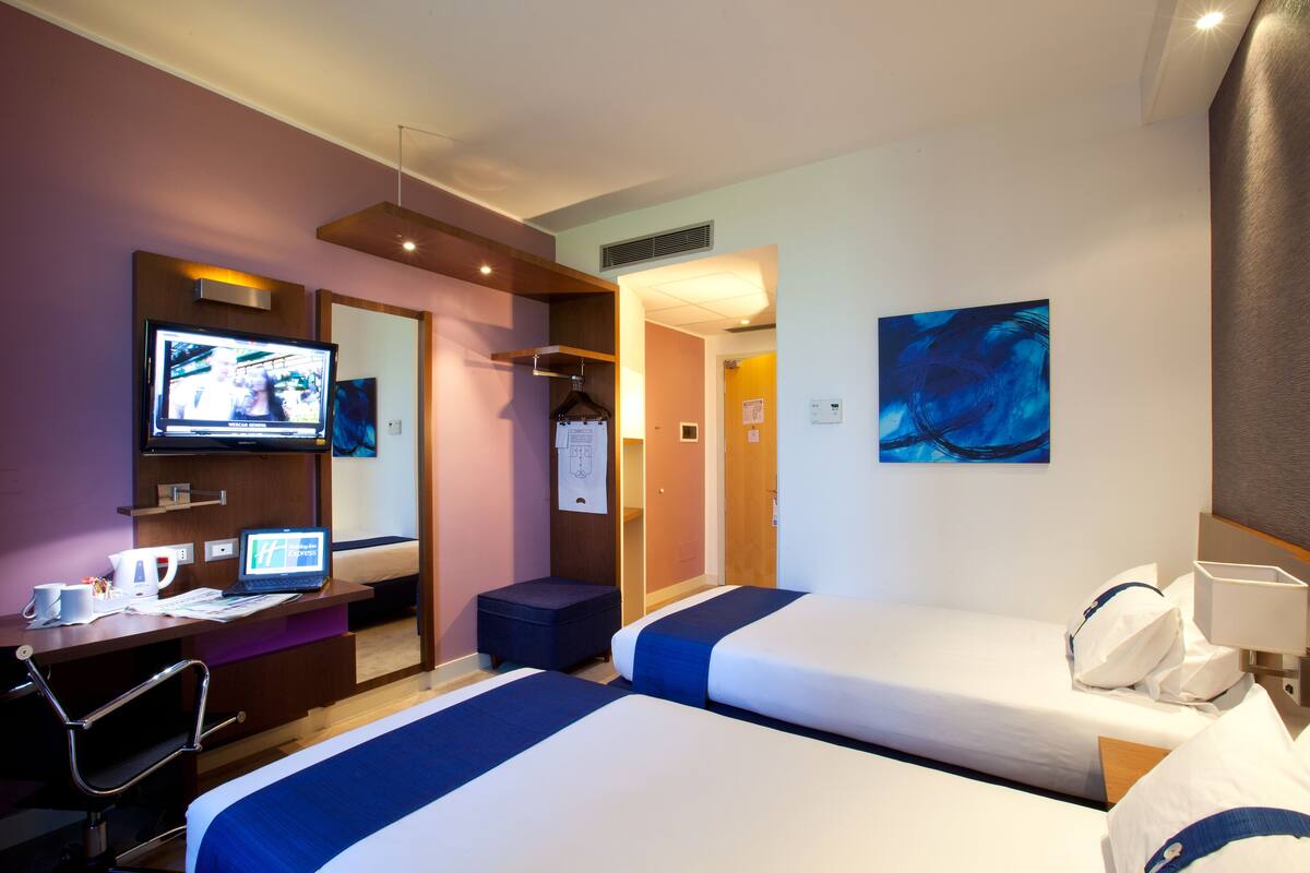 Une chambre d’hôtel avec deux lits, un bureau avec un ordinateur portable, une t élévision et un tableau accroché au mur.