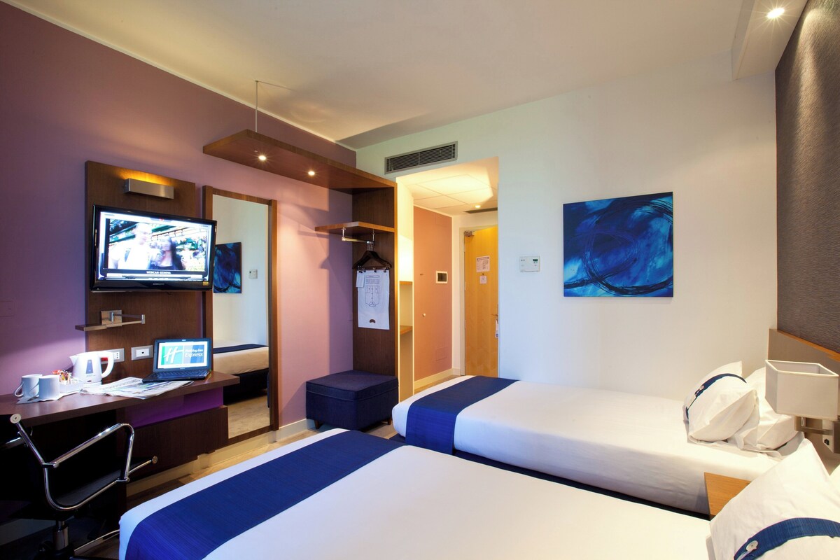 Habitación de hotel con dos camas, un escritorio con una computadora portátil, un televisor y un cuadro en la pared.