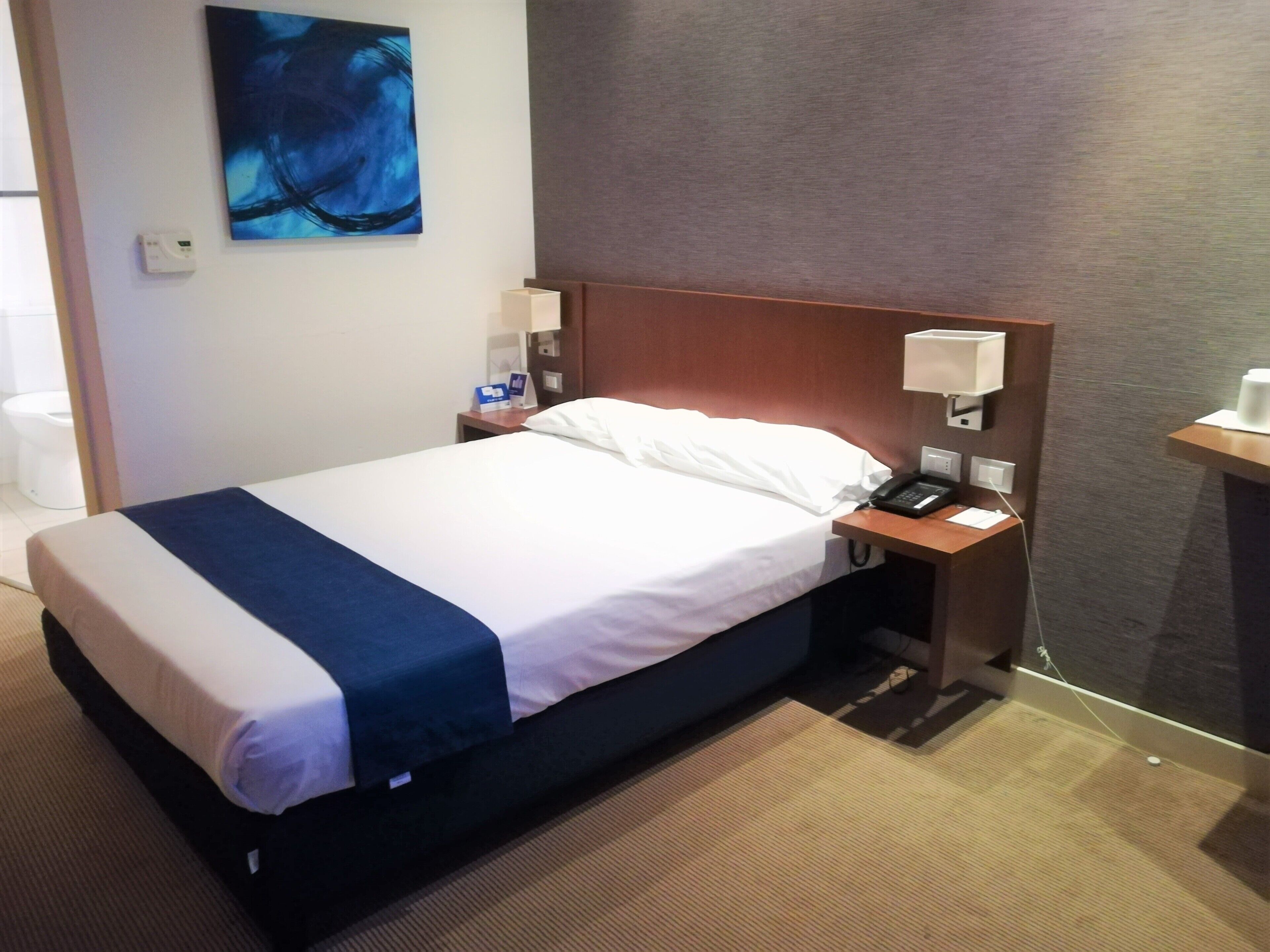 Habitación de hotel con cama, mesita de noche con teléfono, lámpara y un cuadro en la pared.