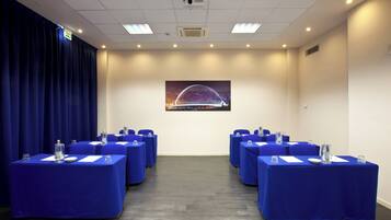 Un salón de conferencias con mesas dispuestas para reuniones, un cartel grande de un puente en la pared y una cortina azul a un lado.
