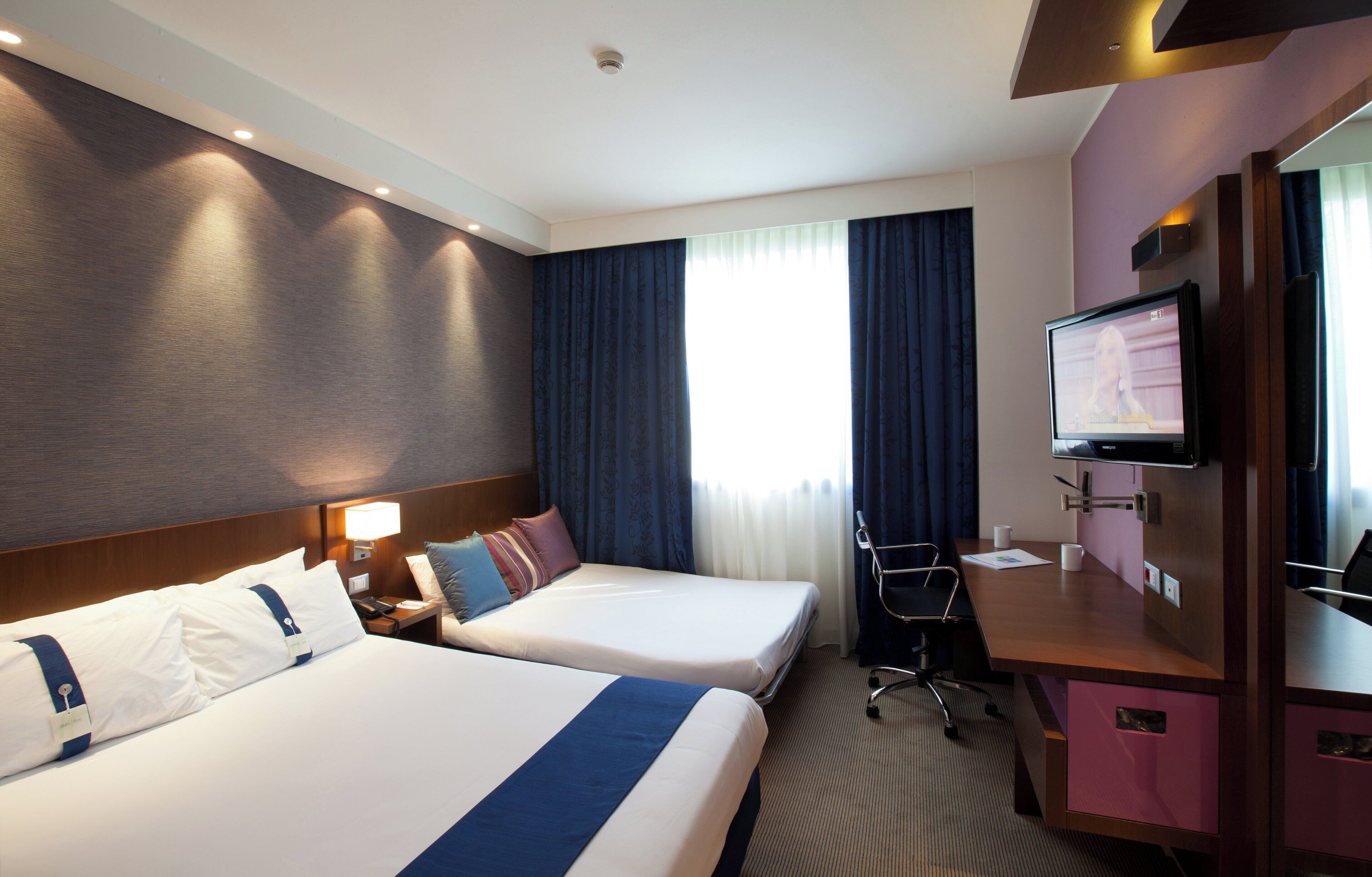 Habitación de hotel con dos camas, un escritorio, un televisor y una ventana con cortinas.