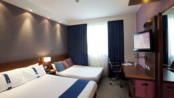 Ein Hotelzimmer mit zwei Betten, einem Schreibtisch, einem Fernseher und einem Vorhangfenster.