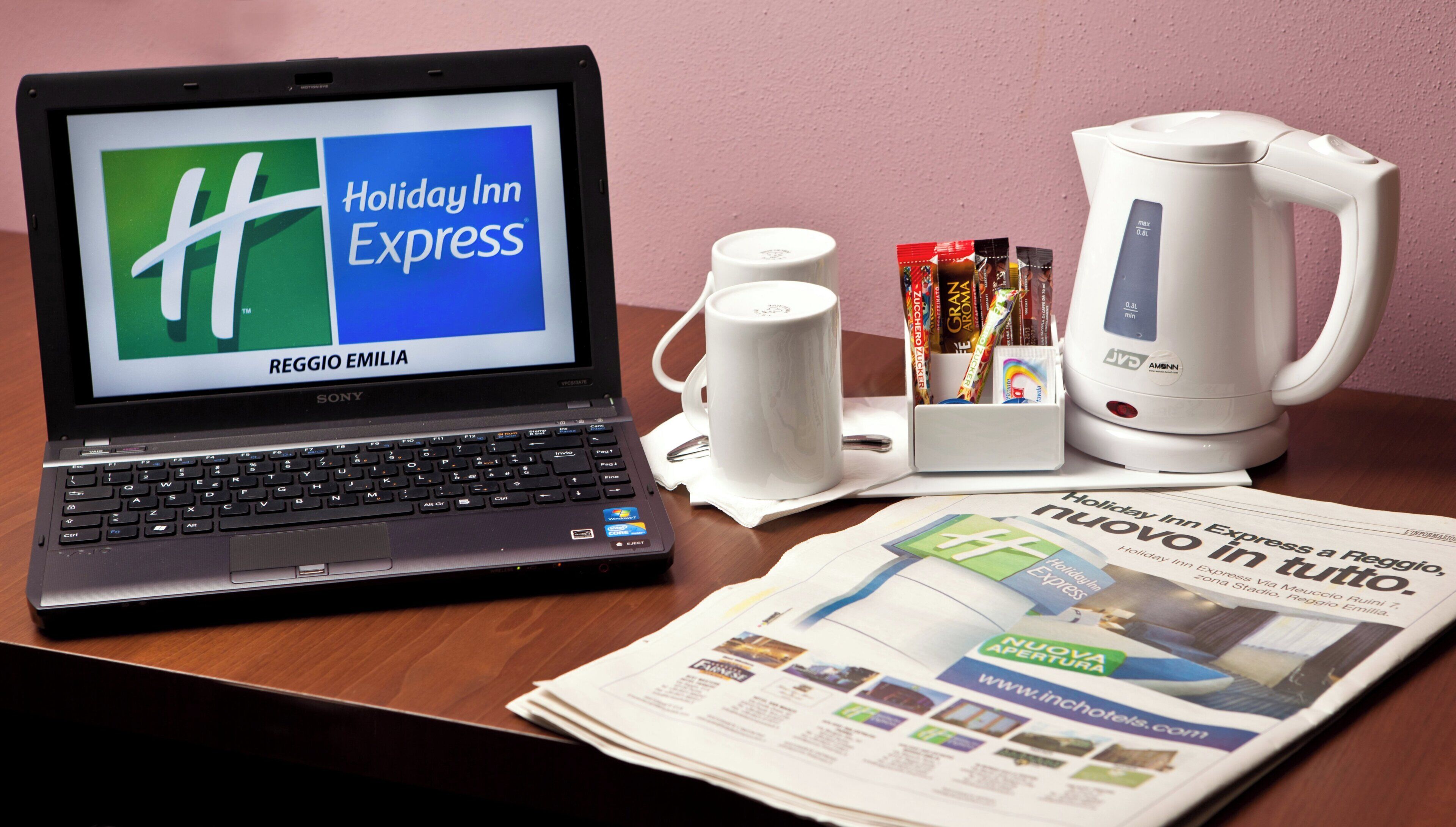 Un ordinateur portable affiche le logo de Holiday Inn Express. Sur une table en bois, on peut également voir une machine à café blanche, une tasse, une cuillère, un paquet de mouchoirs et un journal.