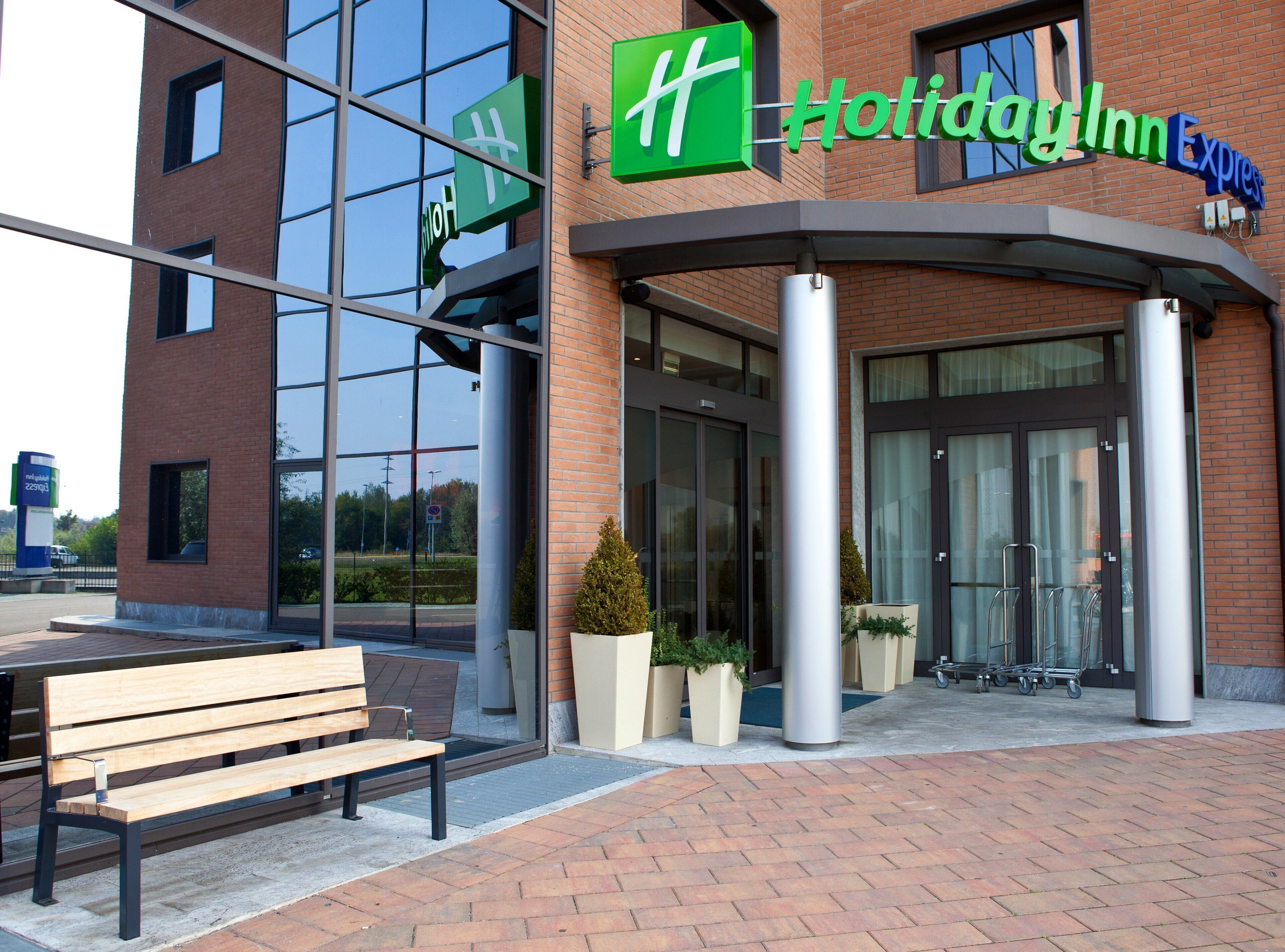 A Holiday Inn Express hotel entrance with a glass facade, a bench outside, and a clear sky.