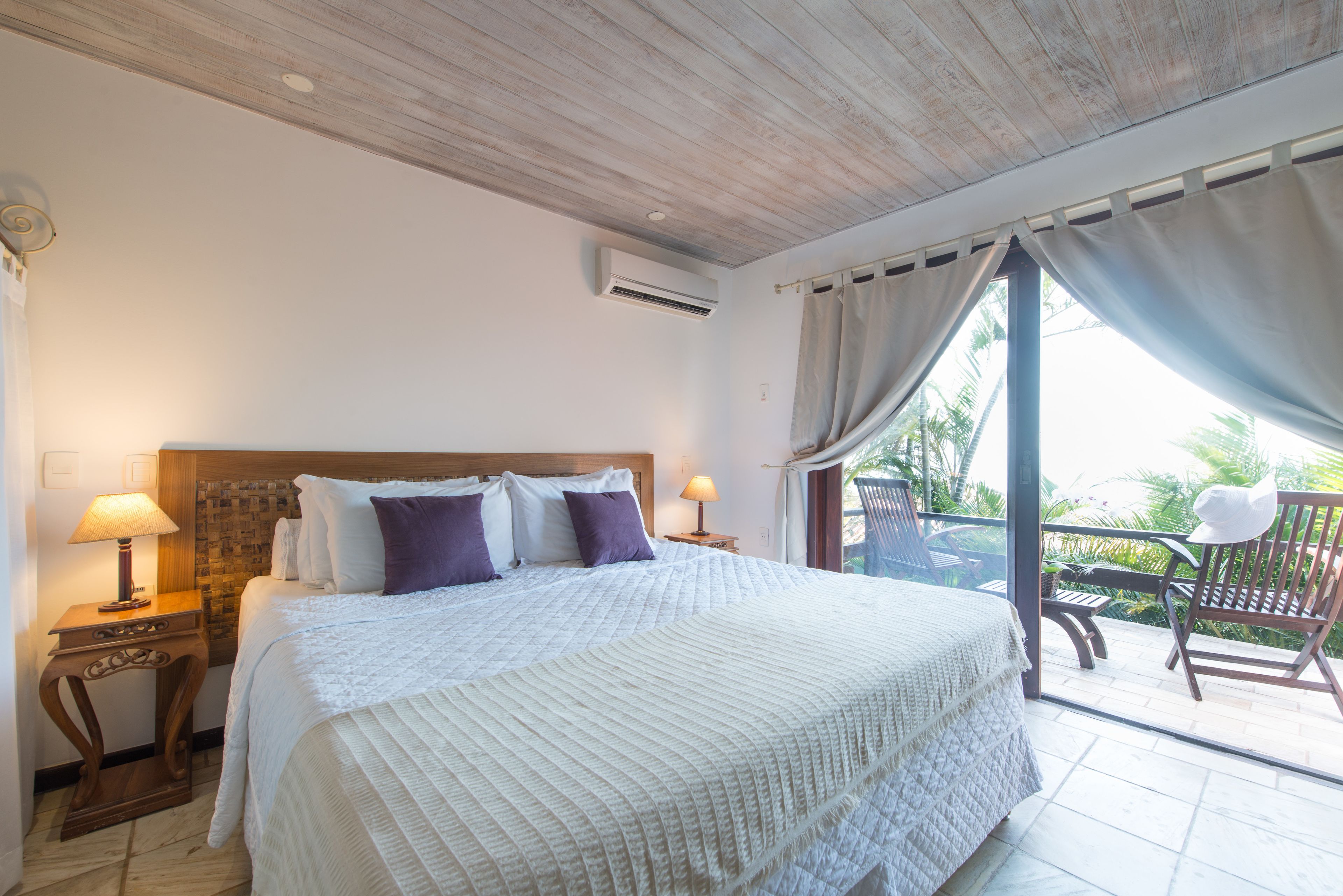 Un dormitorio con una cama grande, dos mesitas de noche con lámparas, un cabecero de madera y vistas al exterior a través de un ventanal corredero.