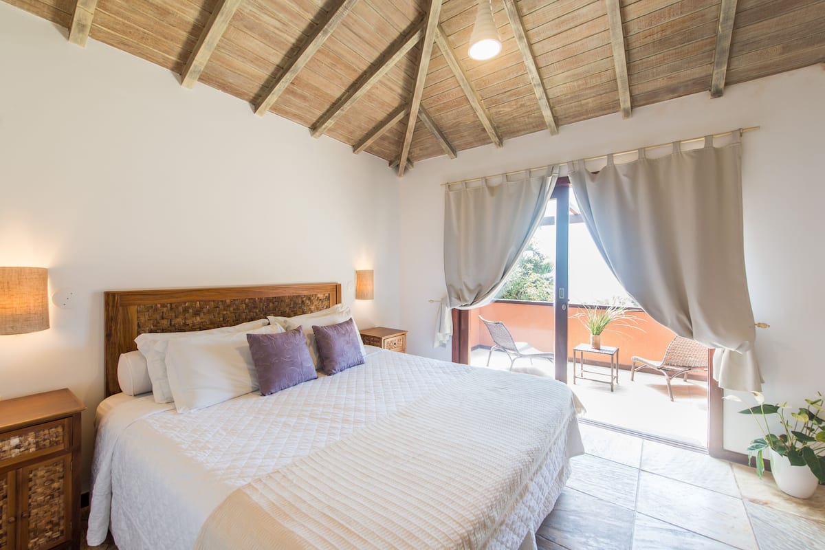A bedroom with a wooden ceiling, a large bed with white bedding, a nightstand, and a view of an outdoor patio with a chair and a table.