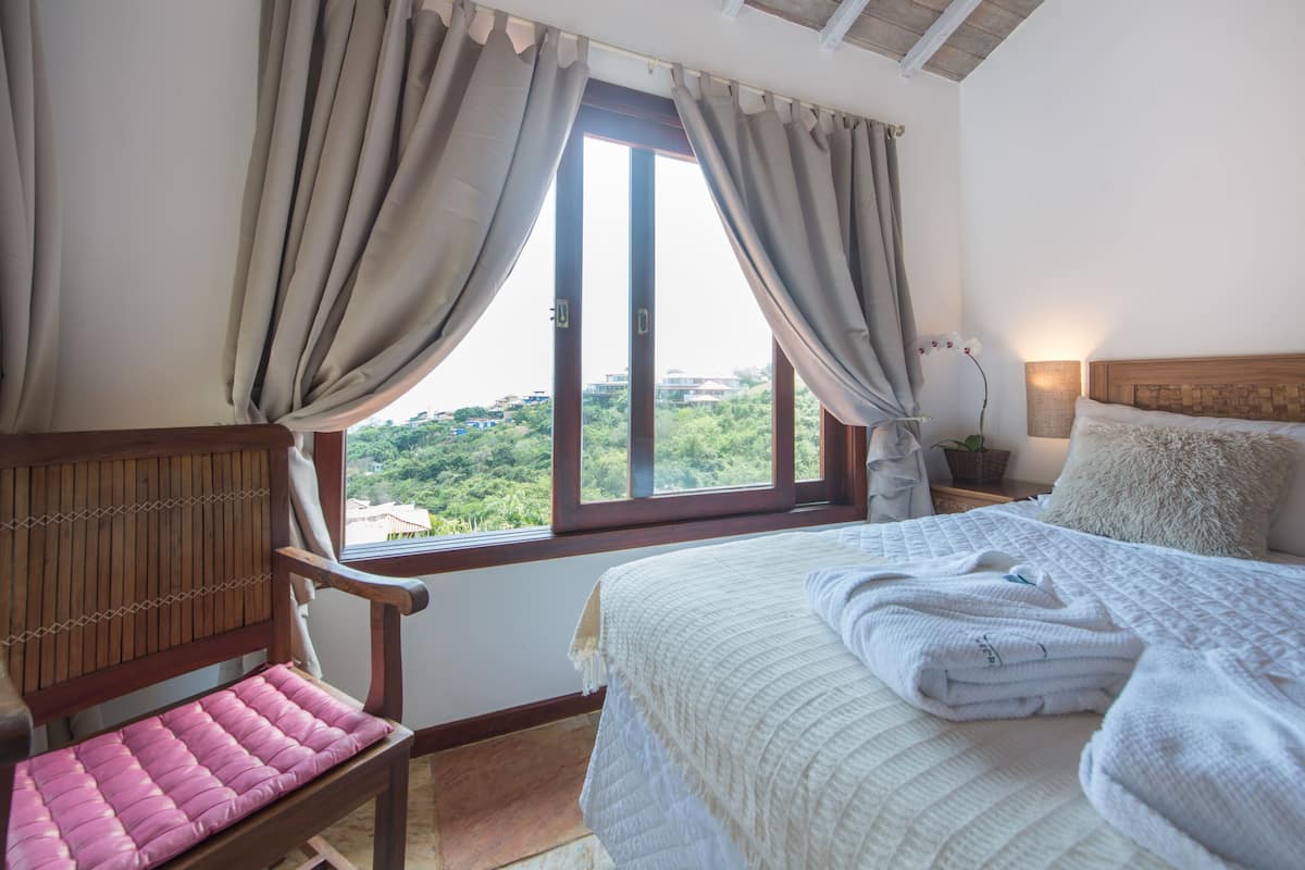 A bedroom with a bed, a chair, a window with curtains, and a view of greenery outside.