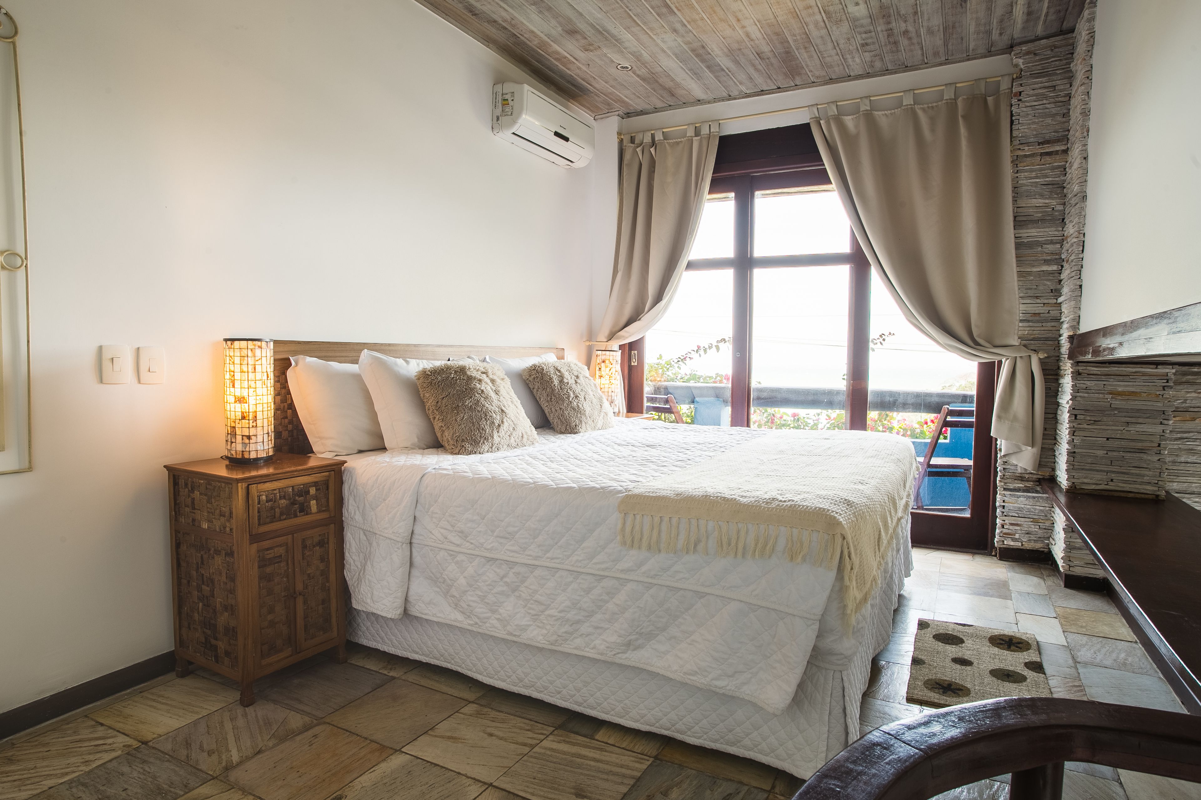 A bedroom with a large bed, a nightstand with a lamp, a window with curtains, and a view of the outside.