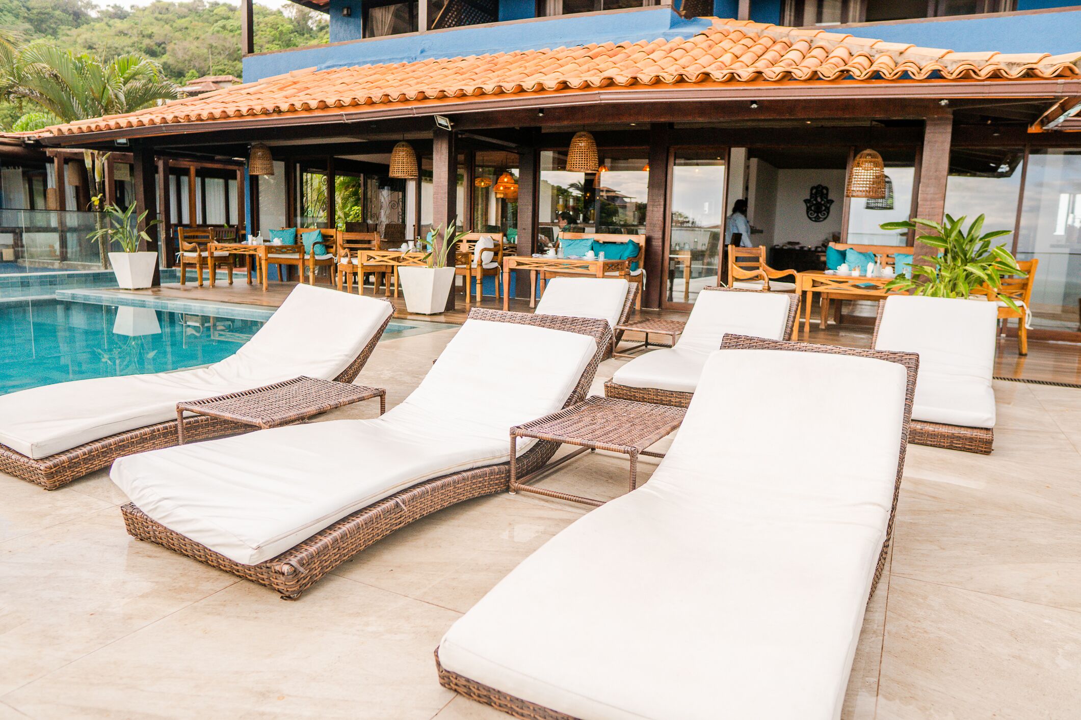 A poolside area with lounge chairs and tables, surrounded by a covered patio with potted plants and a tiled roof.