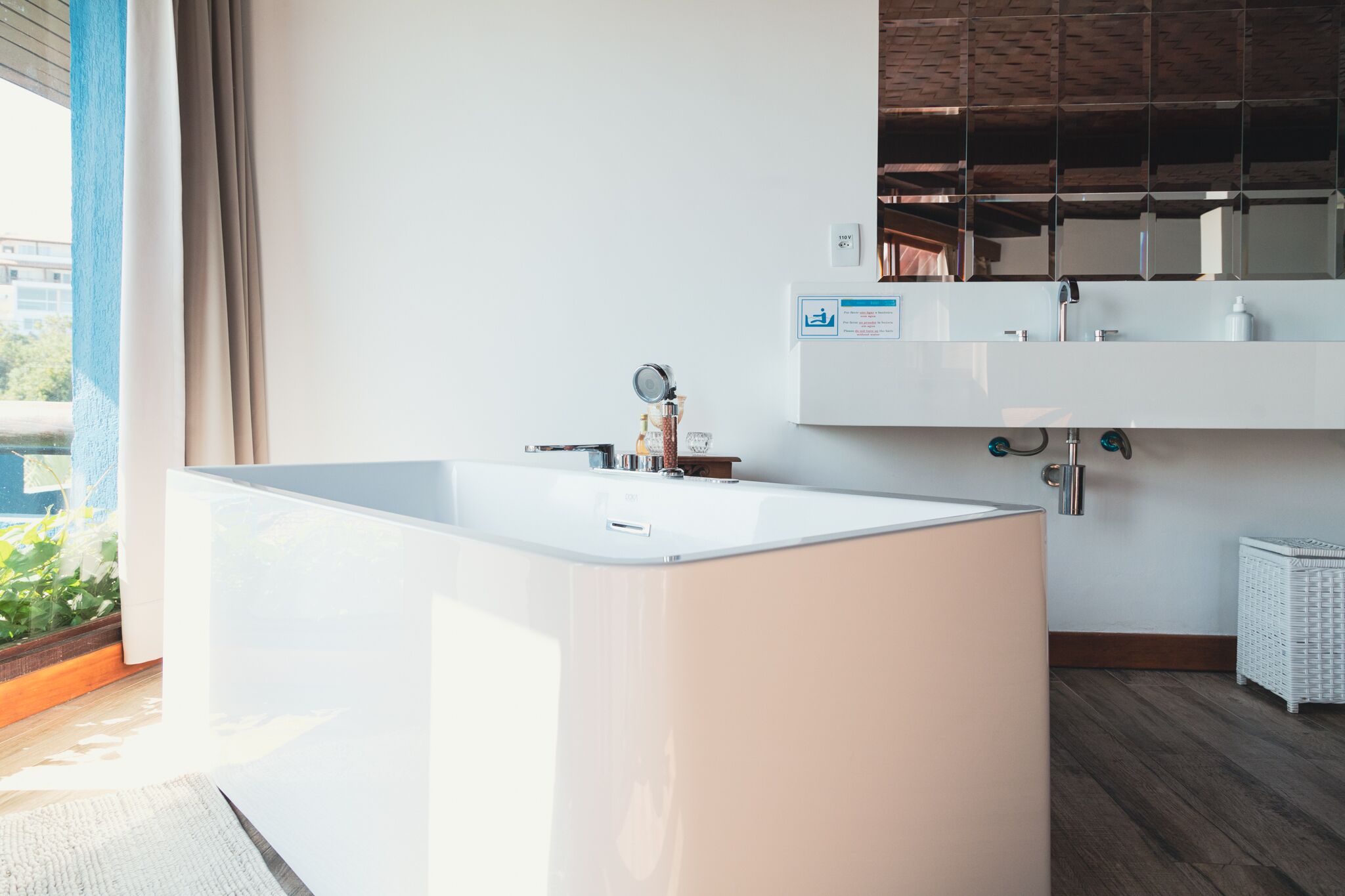A modern bathroom with a large white bathtub, a mirror above the sink, and a view of greenery outside.
