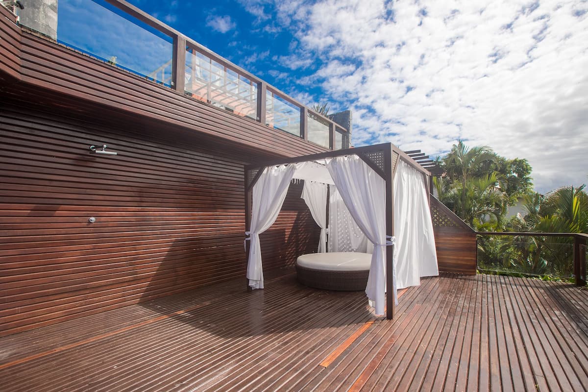 A wooden deck with a canopy bed and a glass balustrade overlooking a tropical landscape.