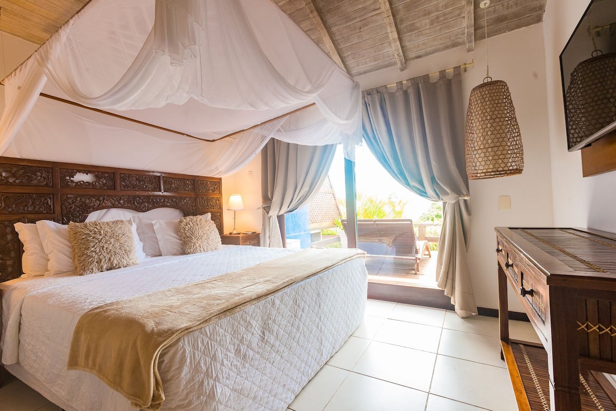 A bedroom with a bed, a wooden headboard, a nightstand, a lamp, a hanging light fixture, and a view of the outdoors.