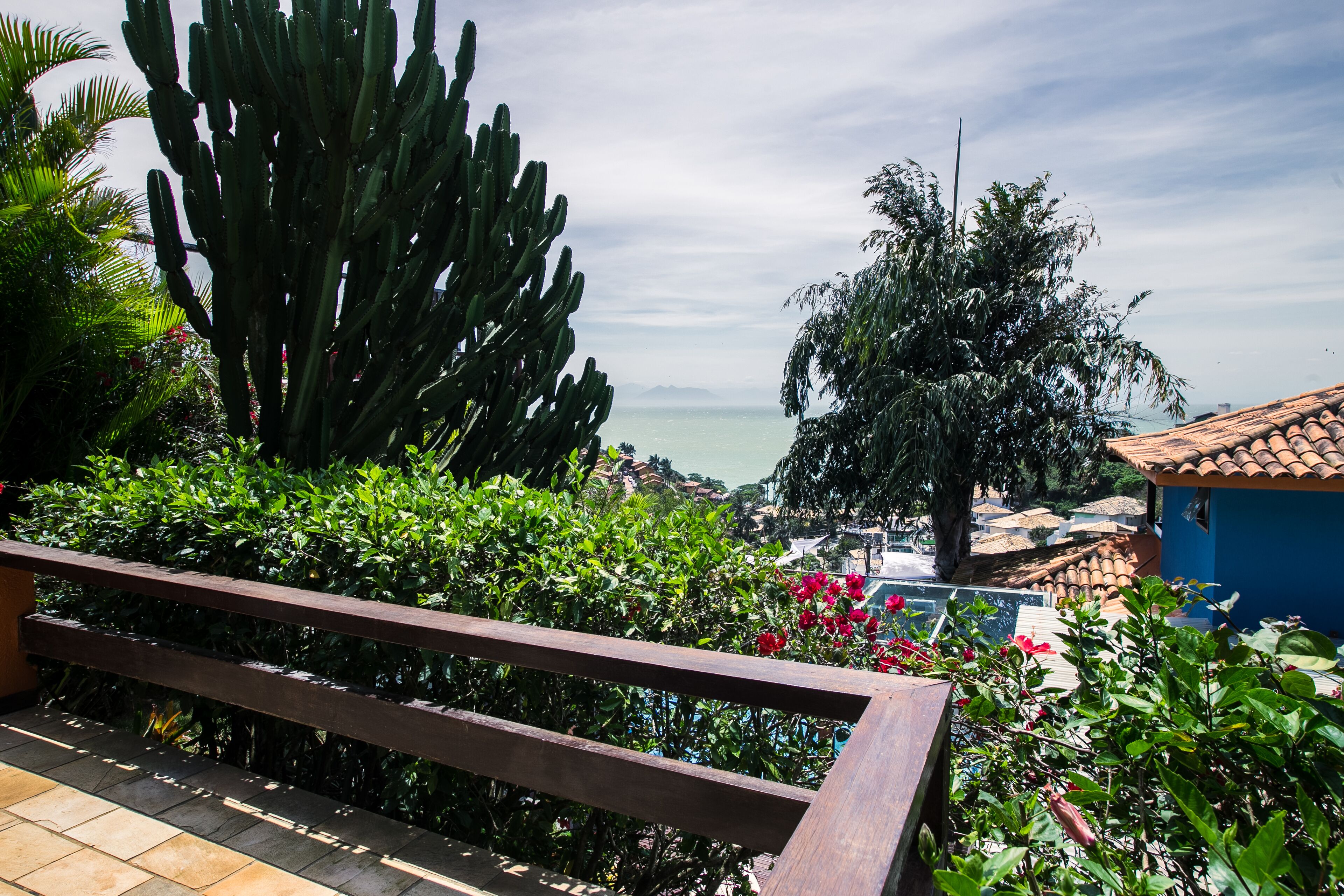 A balcony with a wooden railing overlooking a garden with cacti and flowering plants, a view of the sea, and a blue building with a tiled roof.