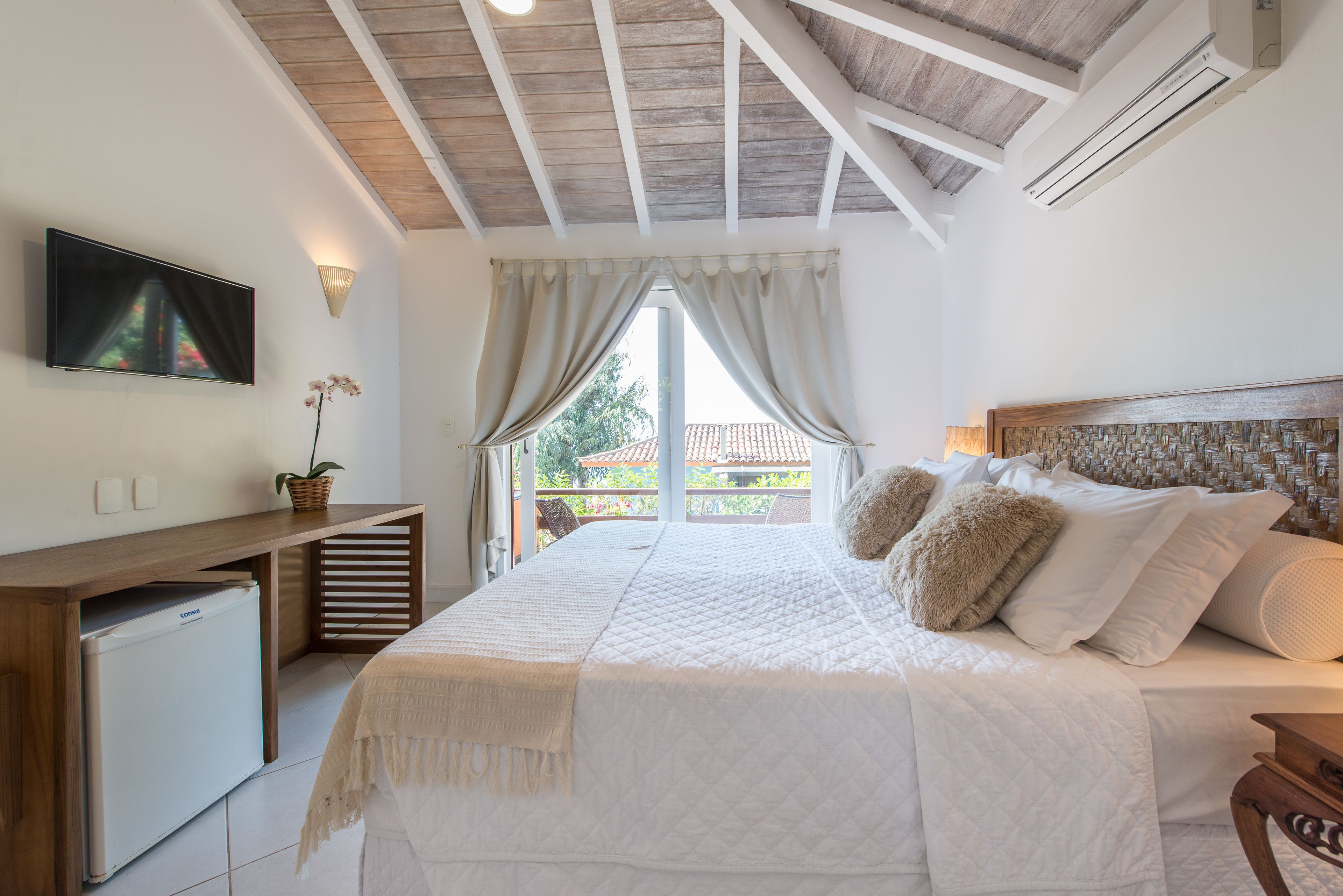 A bedroom with a large bed, a TV mounted on the wall, a small refrigerator, and a view of the outside.