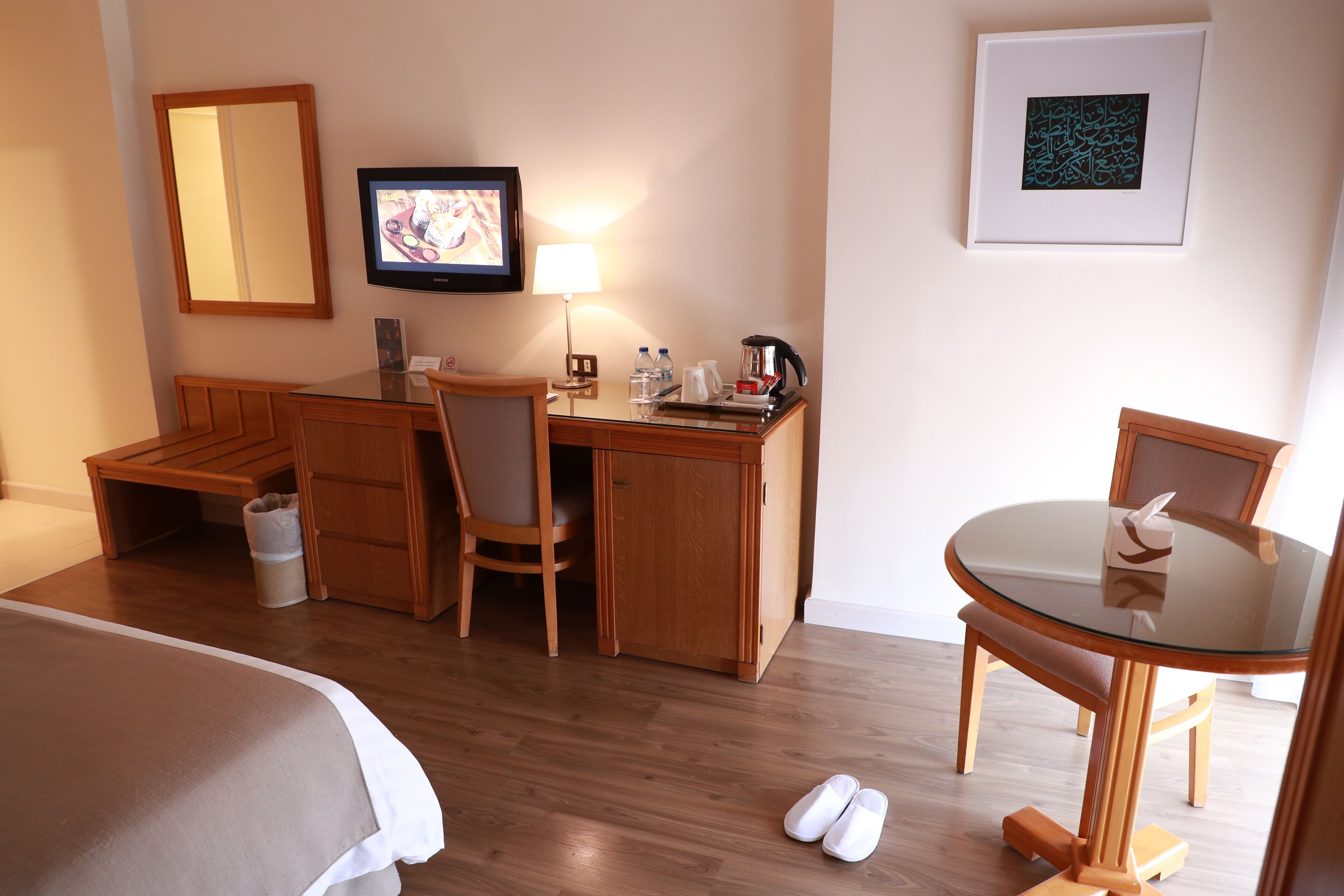 A hotel room with a bed, a desk, a chair, a table, a TV, a lamp, and a framed picture on the wall.