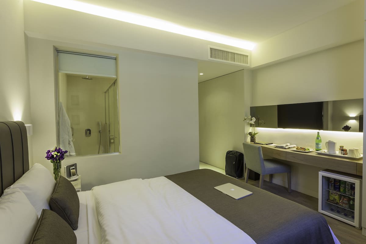 A modern hotel room with a bed, a desk, a chair, a small refrigerator, and a bathroom with a shower and toilet.