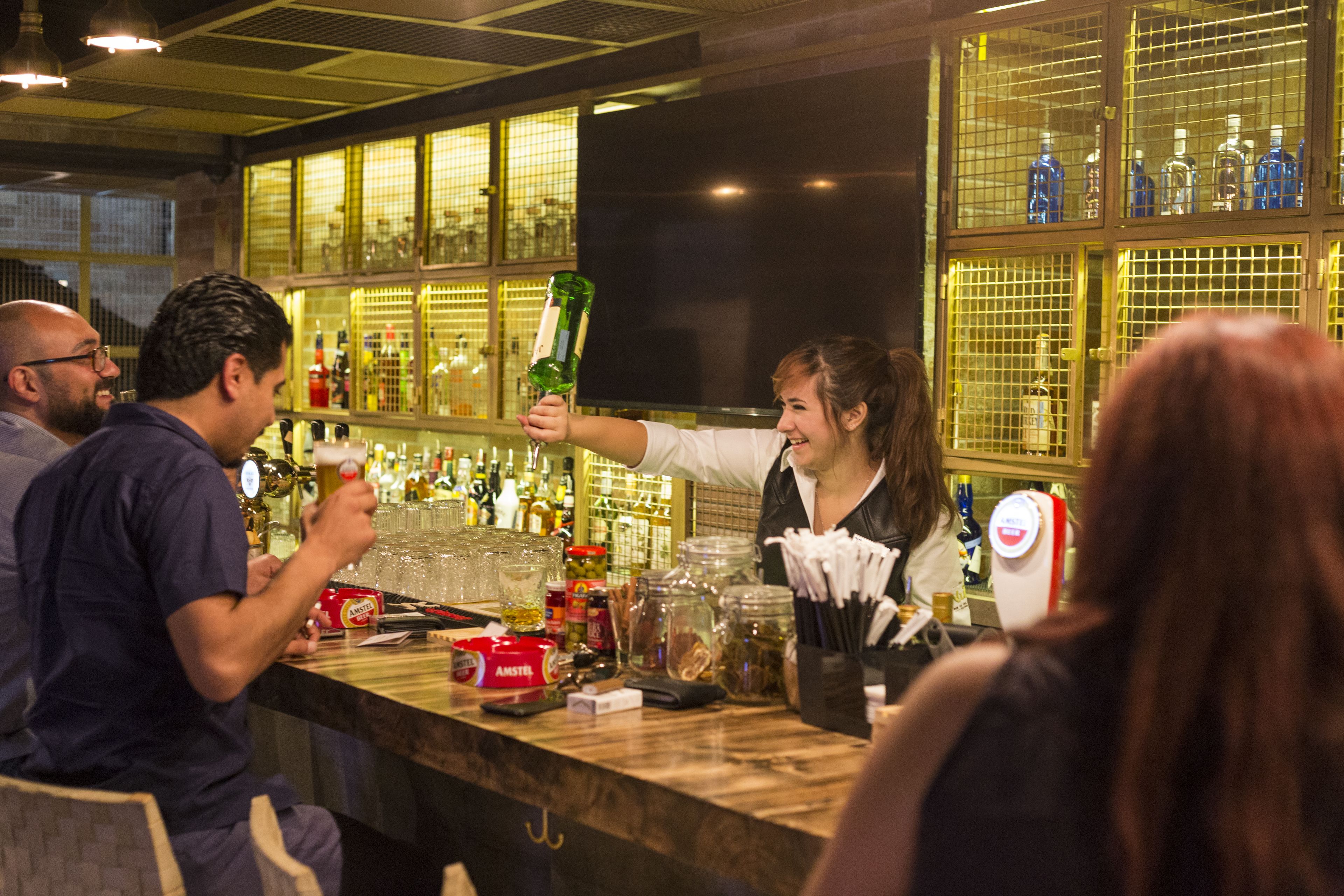 Zona de bar amb estantes plenes de botelles, un taulell amb diversos articles i persones interactuant.
