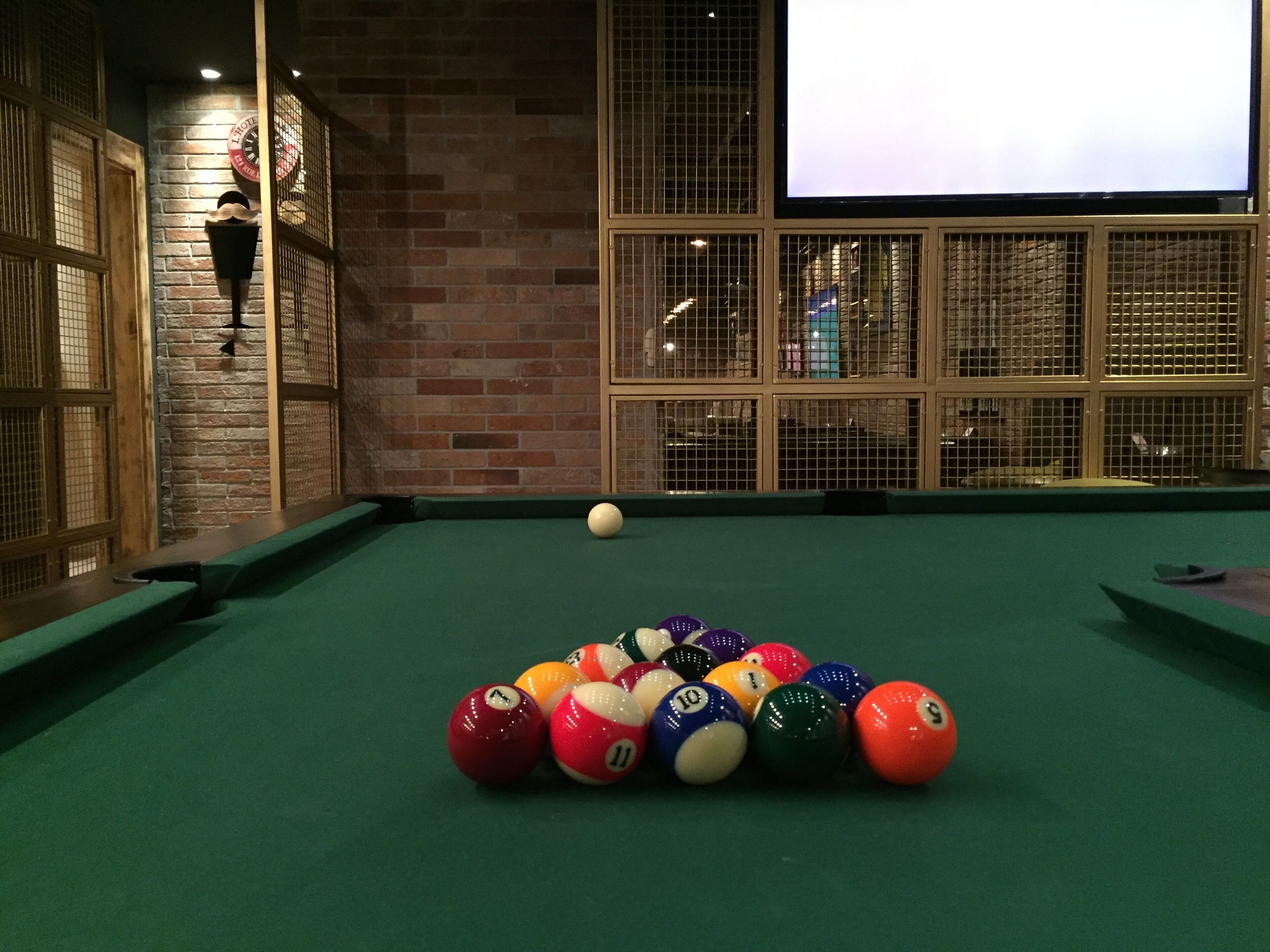 A pool table with a green felt surface, surrounded by a brick wall with a large window and a screen displaying an image.