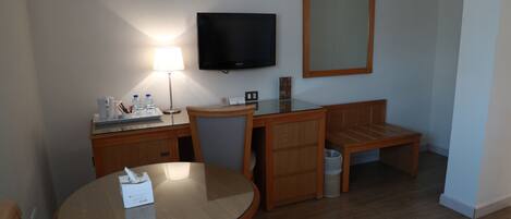 Habitación de hotel con una mesa redonda de vidrio, un escritorio de madera, un televisor montado en la pared, un espejo y un cubo de basura.