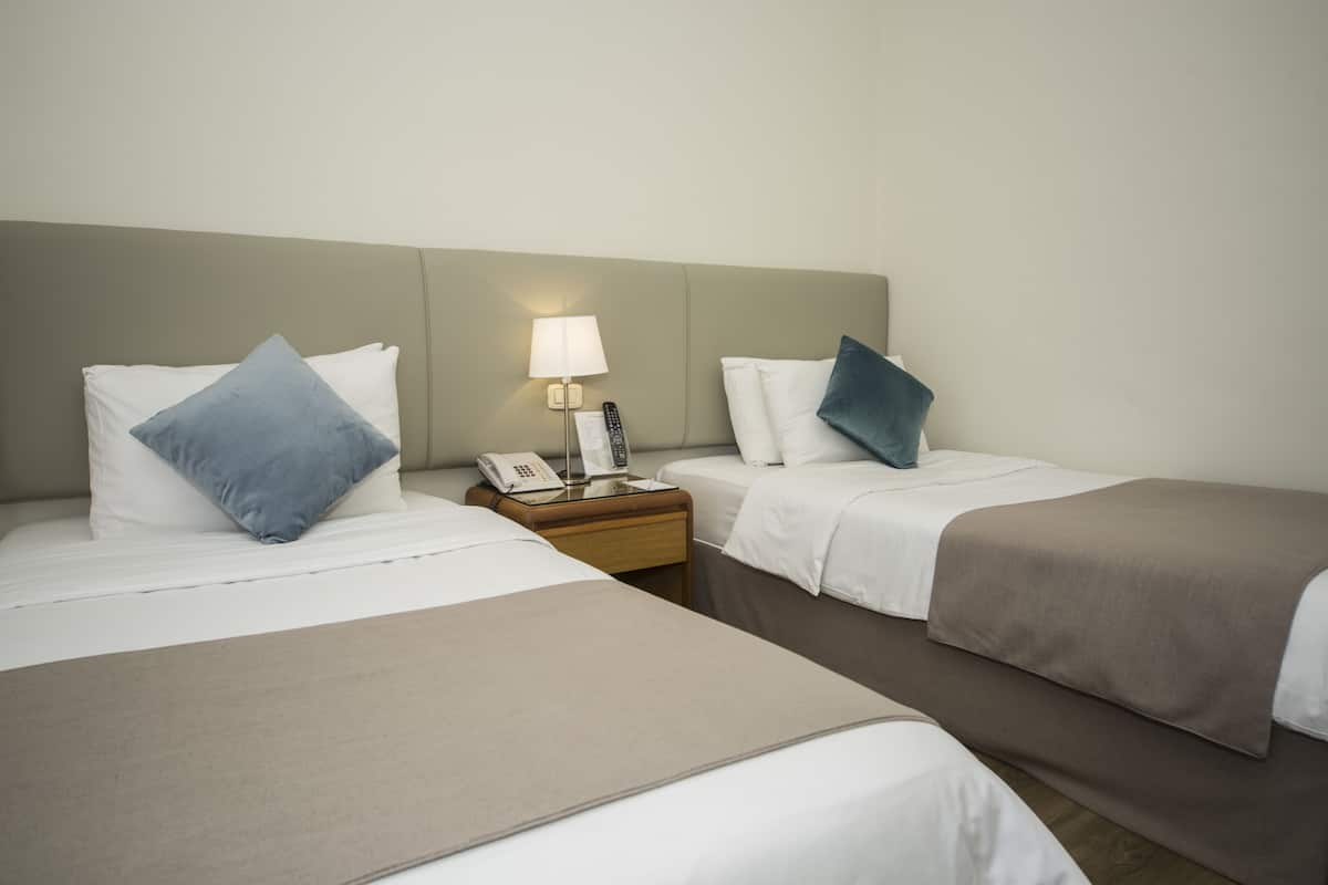 A hotel room with two single beds, a nightstand with a lamp, a phone, and a remote control.