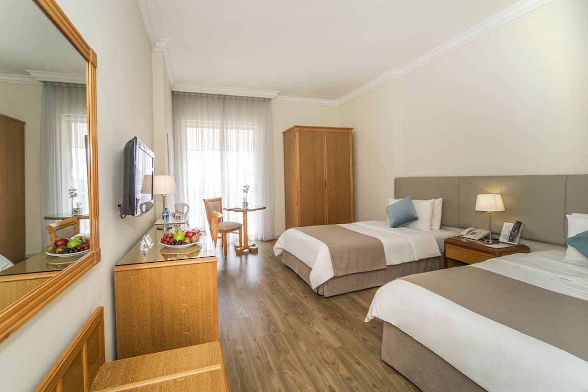 A hotel room with two beds, a desk with a mirror, a small table with a lamp, and a chair.