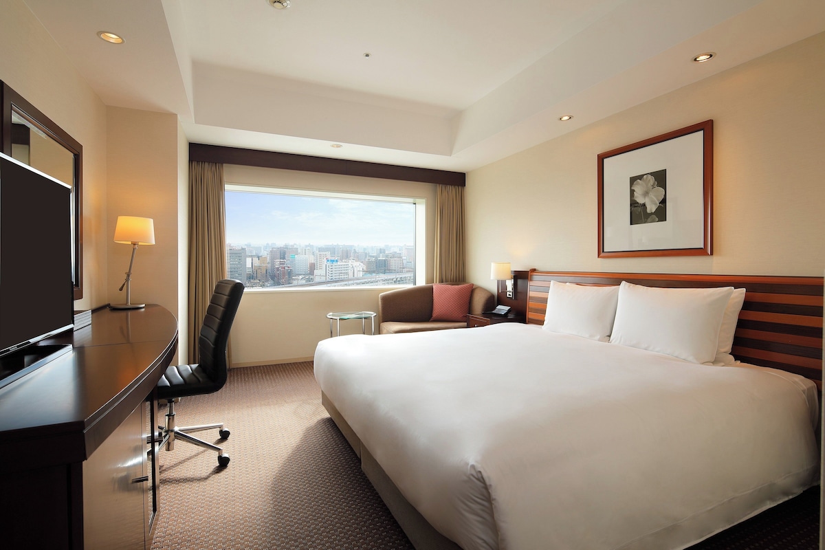Habitación de hotel con una cama grande, un escritorio con una silla, un televisor, una ventana con vistas a la ciudad y un cuadro en la pared.