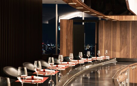 A restaurant with a long counter, multiple tables, and a city view.