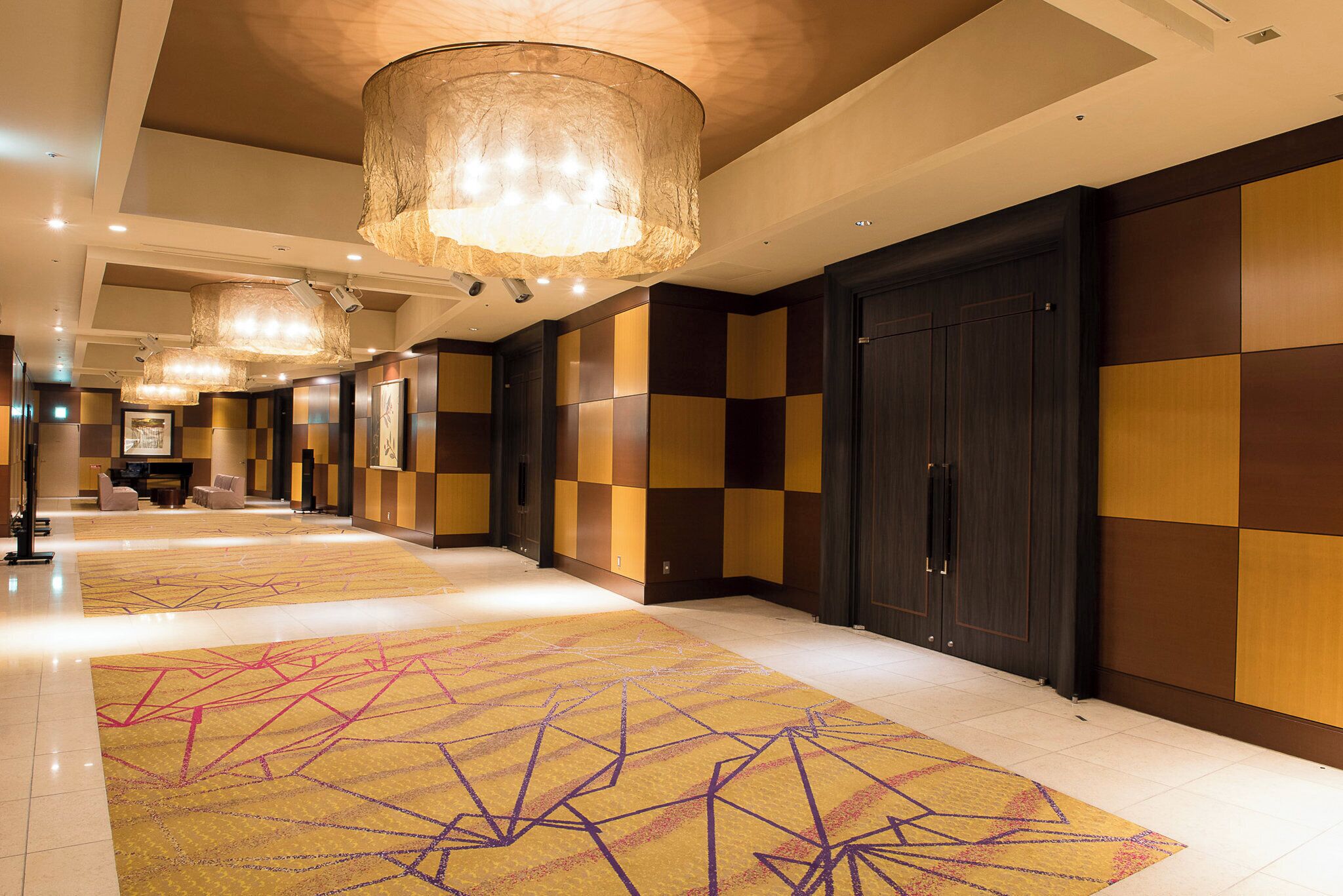 A hotel corridor with a checkered wall pattern, a patterned carpet, and a large chandelier.