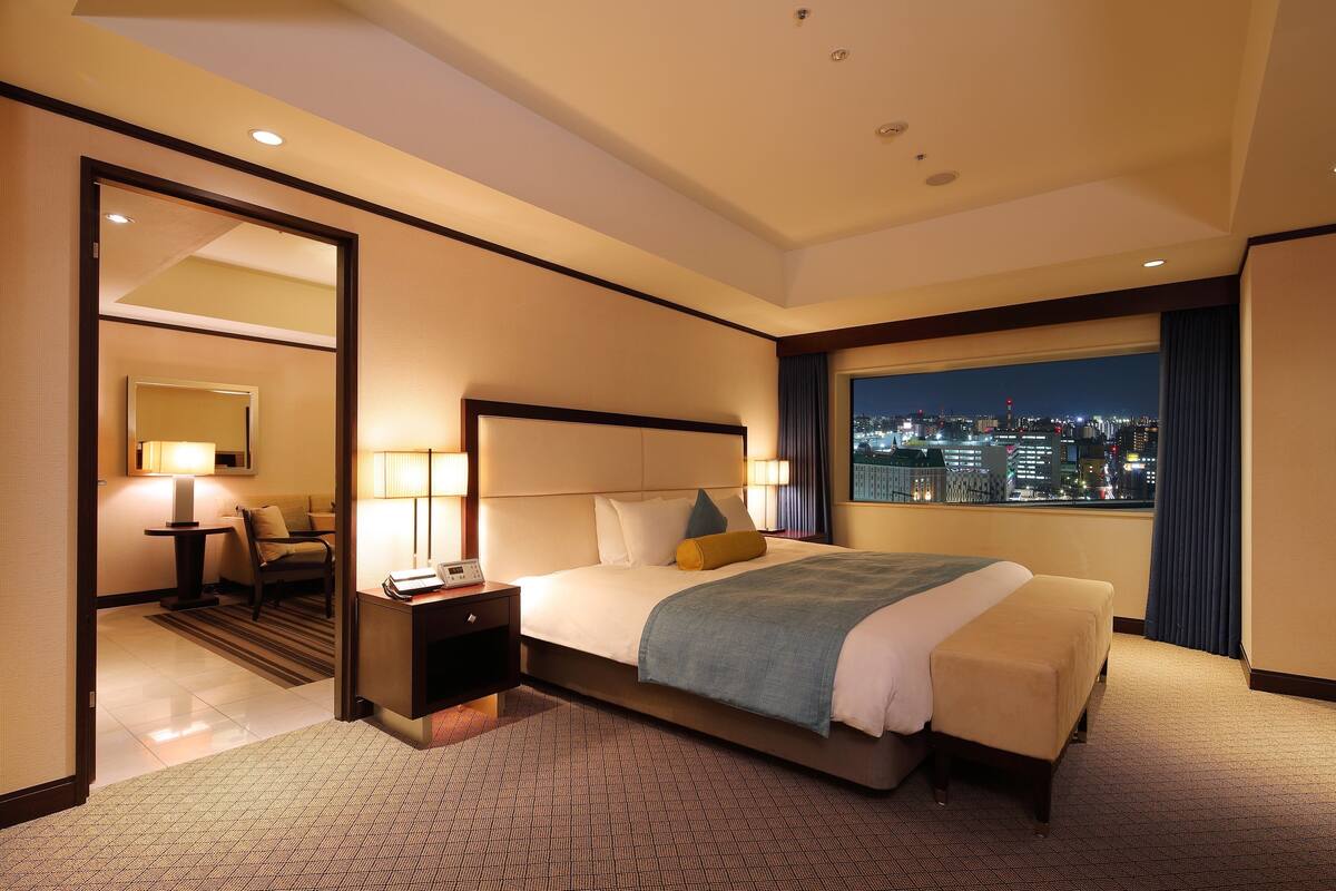 Une chambre d’hôtel avec un lit, des lampes de chevet, un bureau et une vue sur le paysage urbain illuminé la nuit.