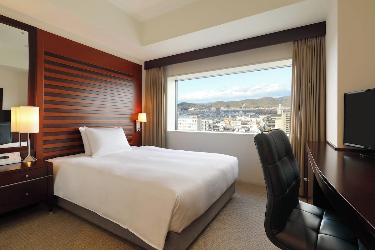 Une chambre d’hôtel avec un grand lit, un bureau, une chaise, une télévision, une fenêtre donnant sur la ville et une tête de lit en bois.