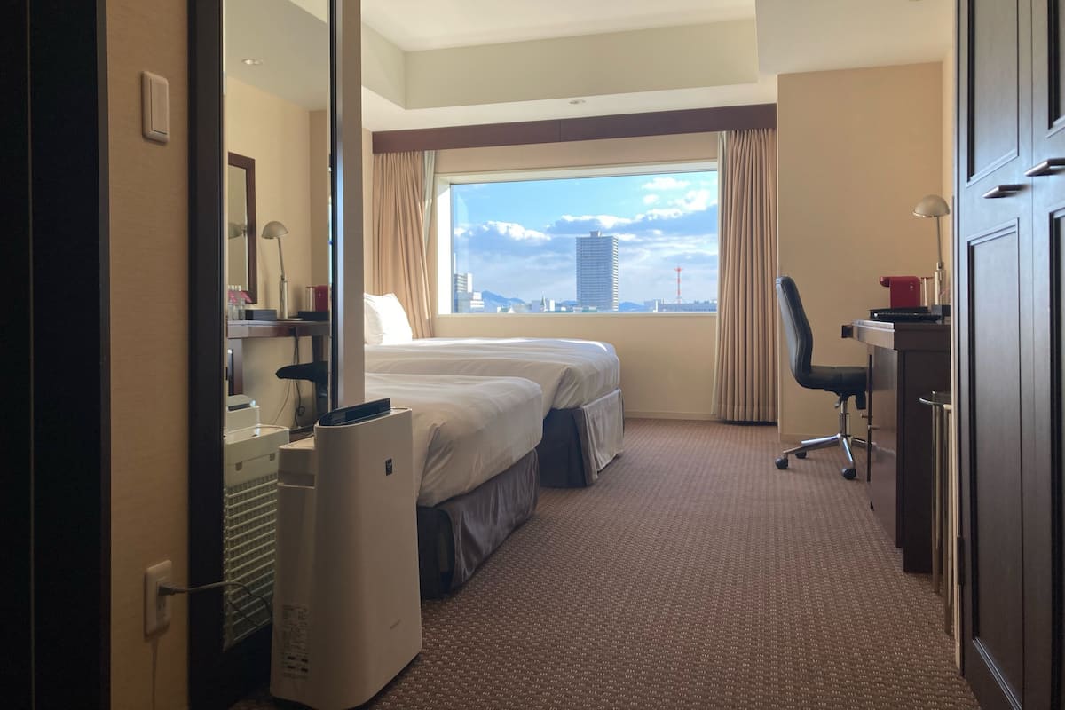 A hotel room with a bed, a desk, a chair, and a large window with a city view.