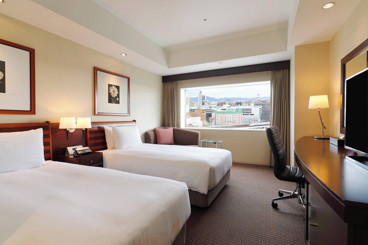 Une chambre d’hôtel avec deux lits, un bureau, une chaise, une télévision et une fenêtre donnant sur la ville.