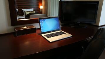 A hotel room with a wooden desk, a laptop, a television, and a bedside table with a lamp.