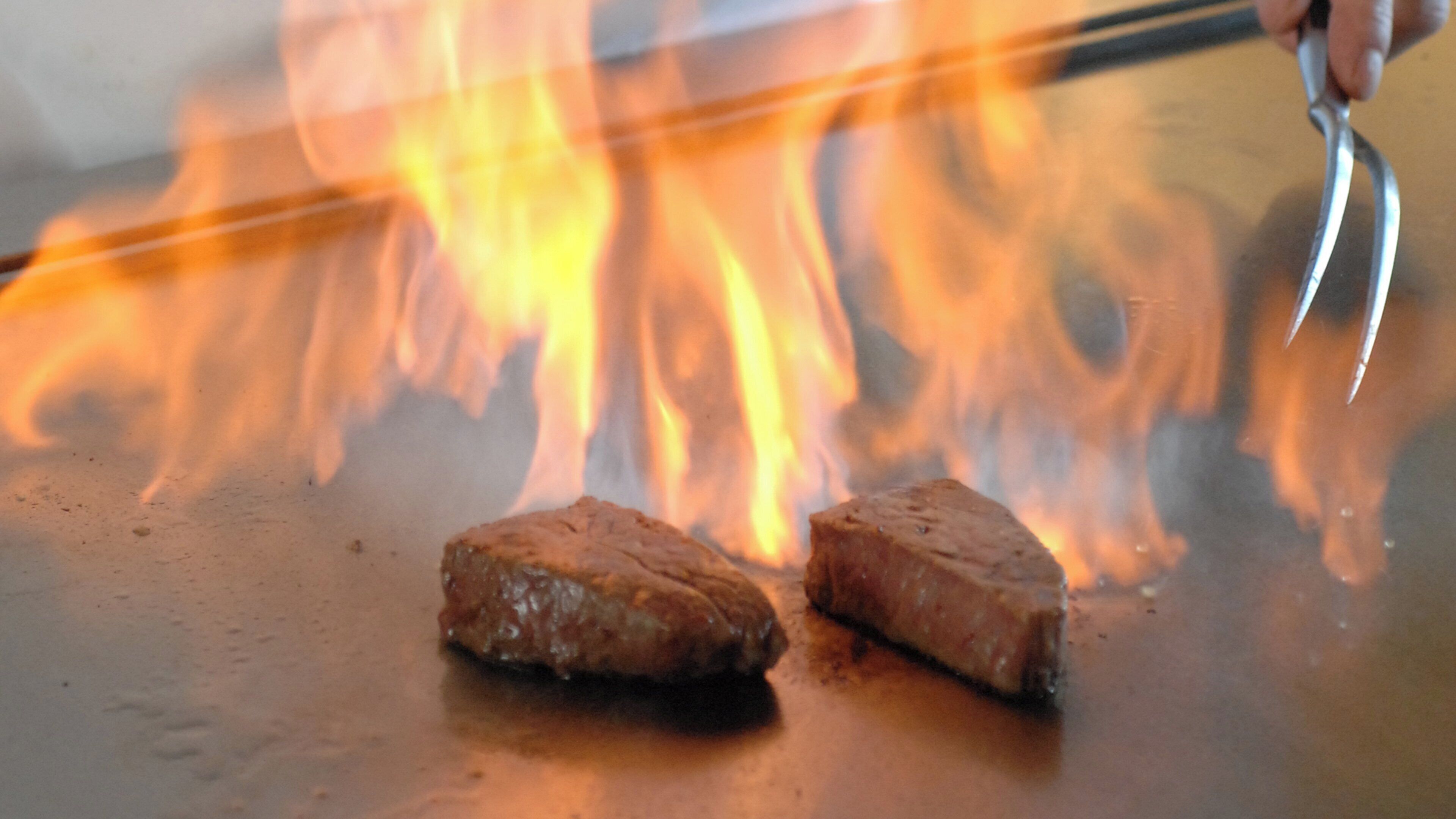 Two pieces of meat being grilled on a flat surface with flames in the background.