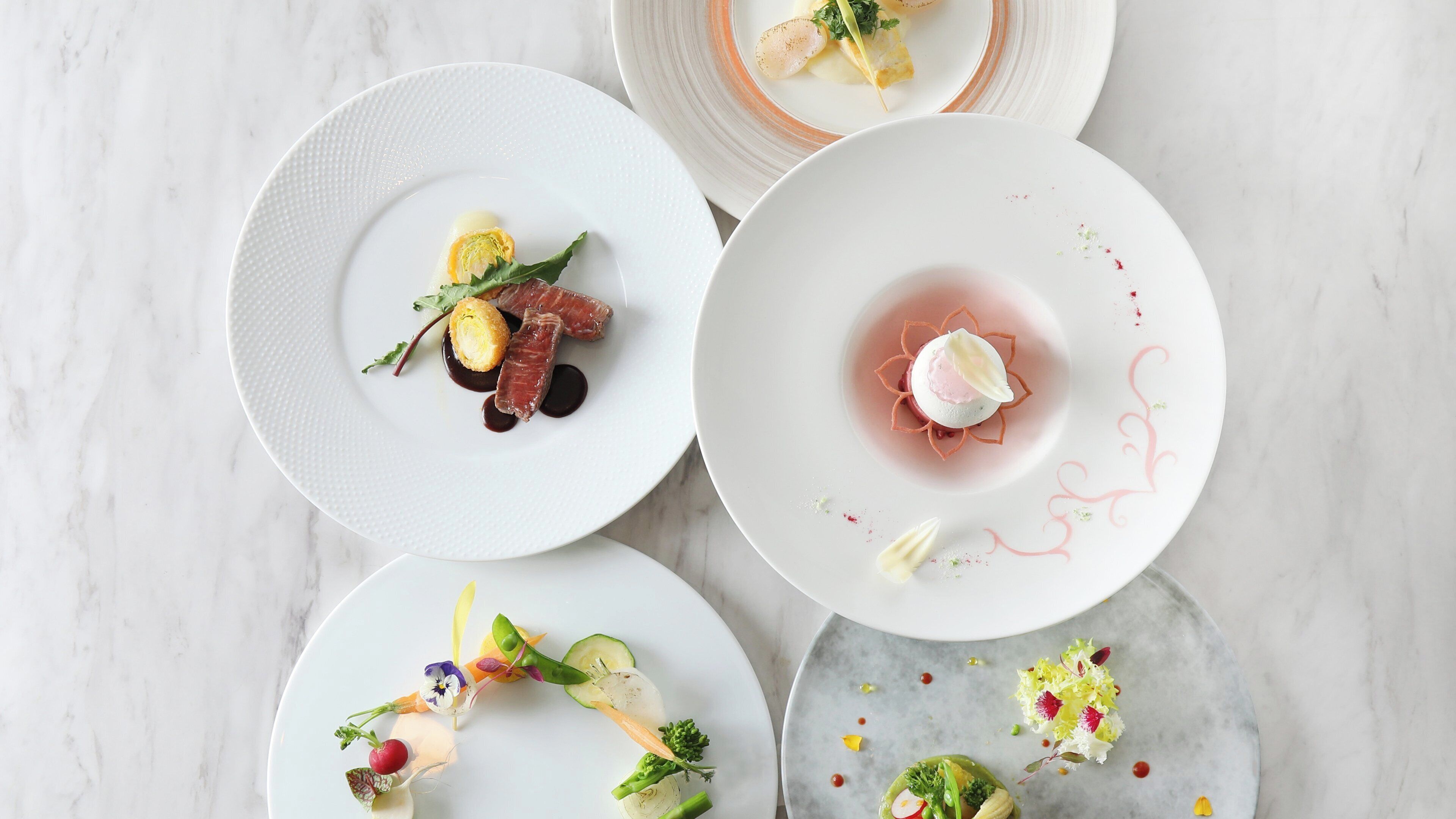 Four plates of food with various dishes on a marble surface.