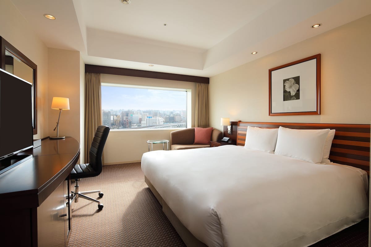 Une chambre d’hôtel avec un grand lit, un bureau avec une chaise, une tél évision, une fenêtre donnant sur la ville et un tableau encadré au mur.