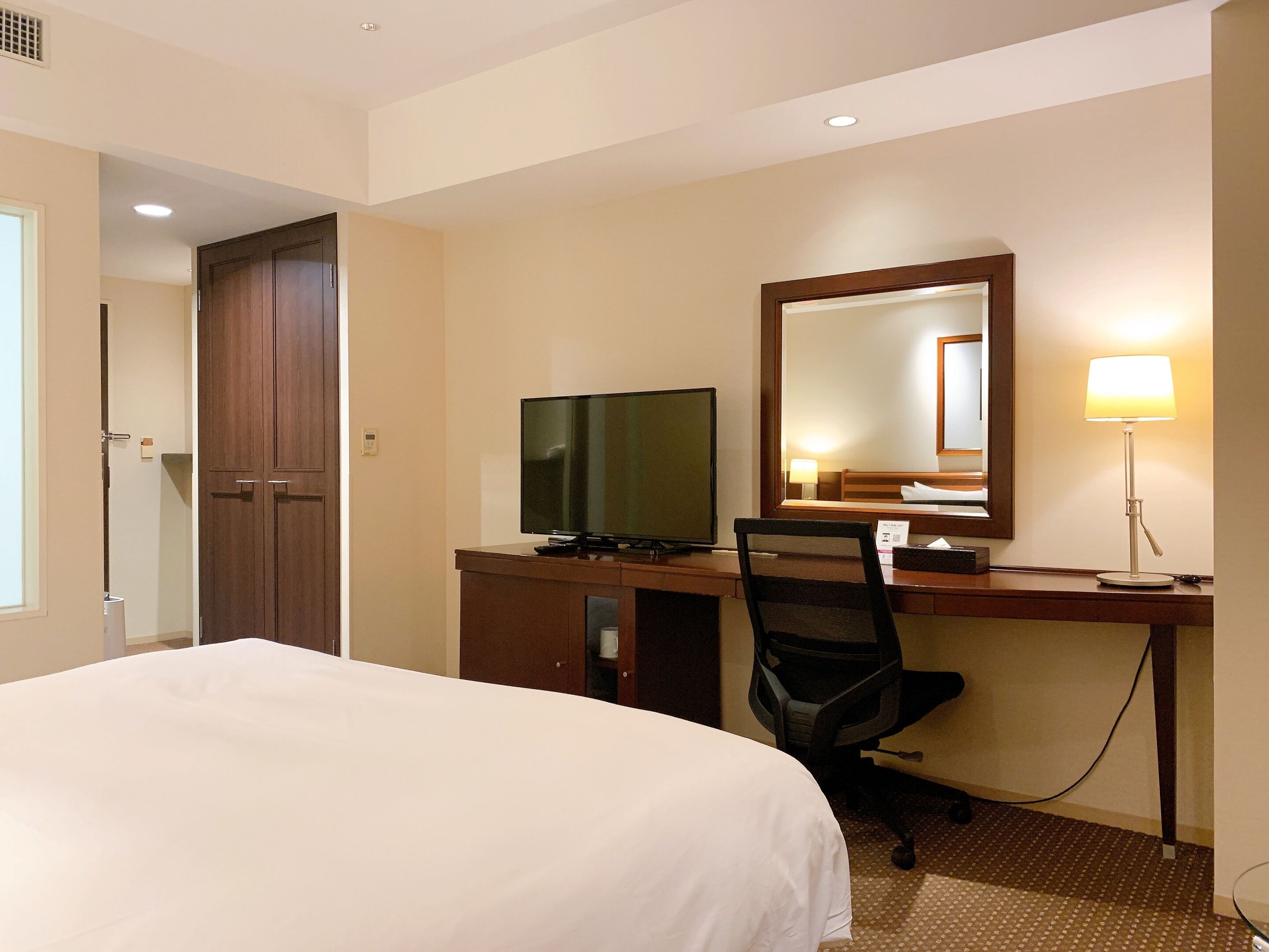 Ein Hotelzimmer mit einem Bett, einem Schreibtisch mit Fernseher, einem Sessel, einer Schreibtischlampe und einem großen Spiegel.