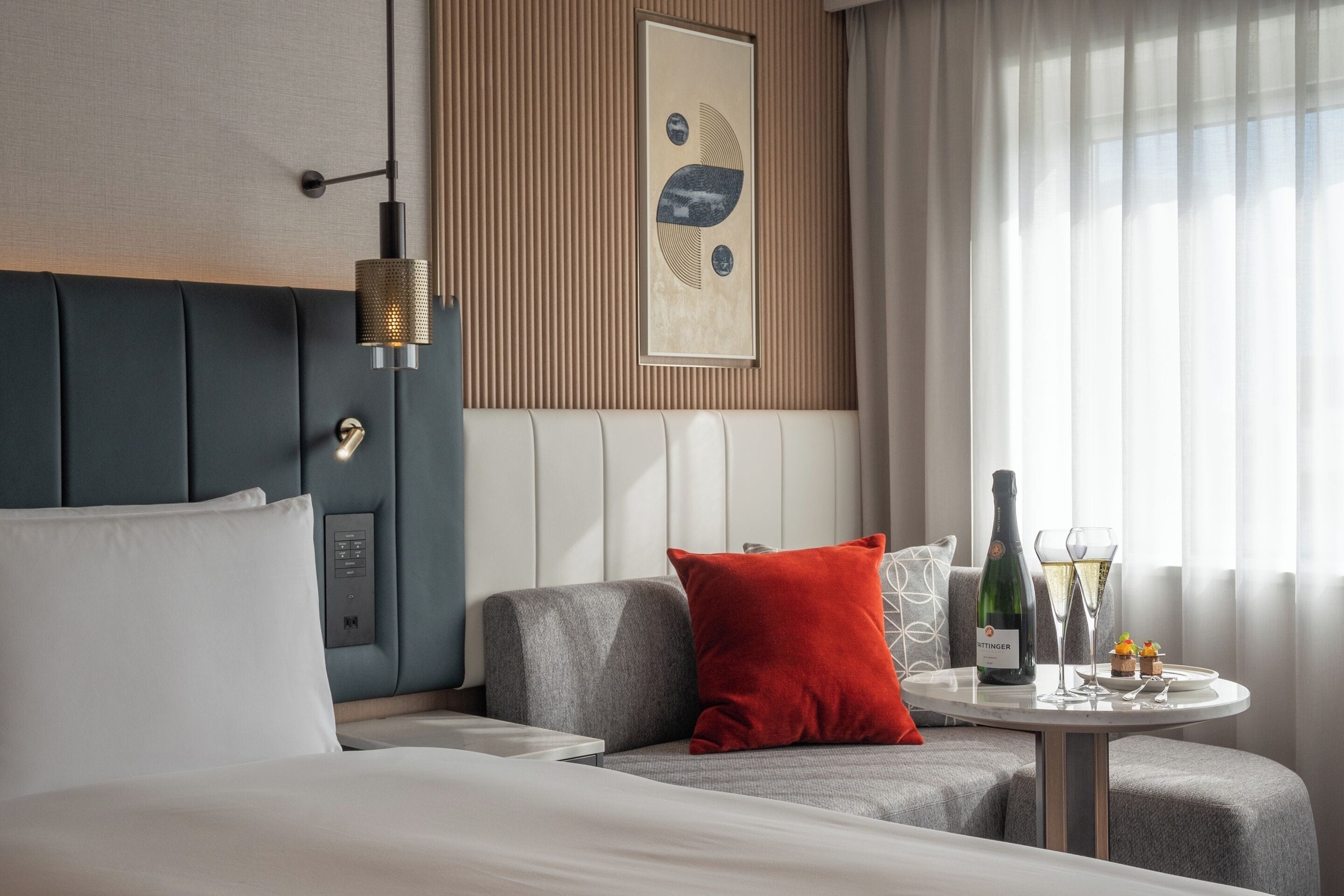 Una habitación de hotel moderna con una cama, un sofá, una mesita con una botella y vasos, y una ventana con cortinas.