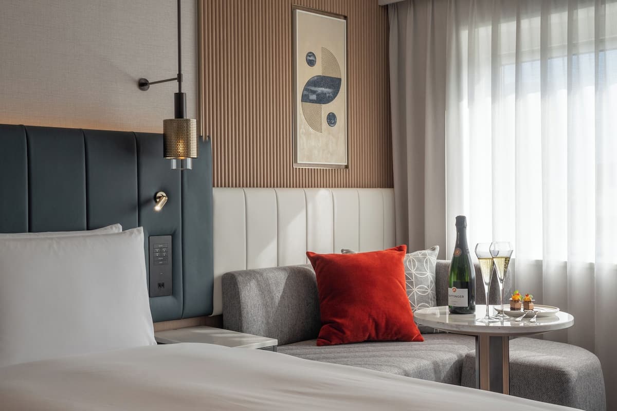 A modern hotel room with a bed, a sofa, a small table with a bottle and glasses, and a window with curtains.