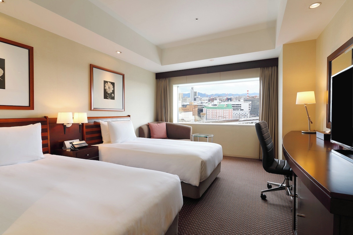 Habitación de hotel con dos camas, un escritorio, una silla, un televisor y una ventana con vista a la ciudad.