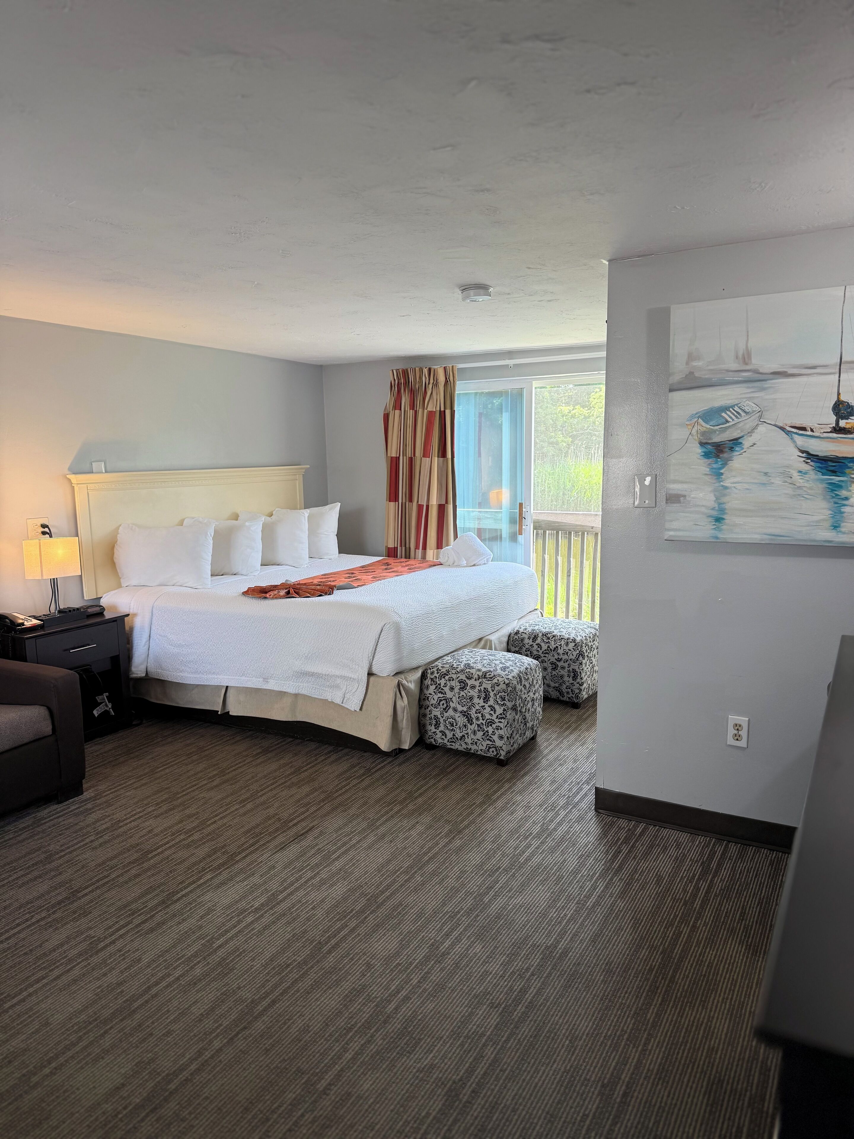 Habitación de hotel con cama, mesita de noche, lámpara y un cuadro de un barco en la pared.