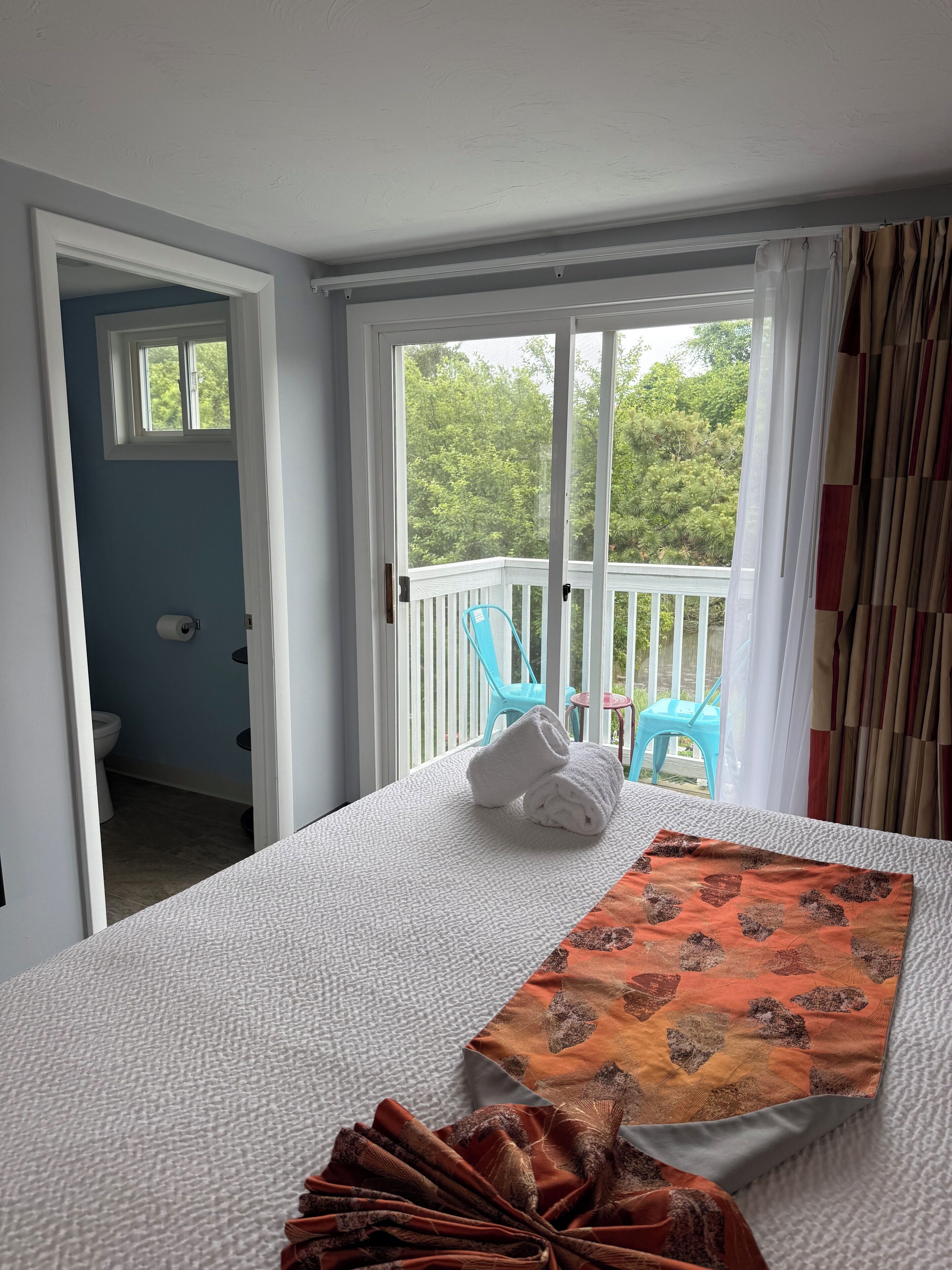 Quarto de hotel com varanda, uma cama com colcha branca e laranja e uma cadeira azul.
