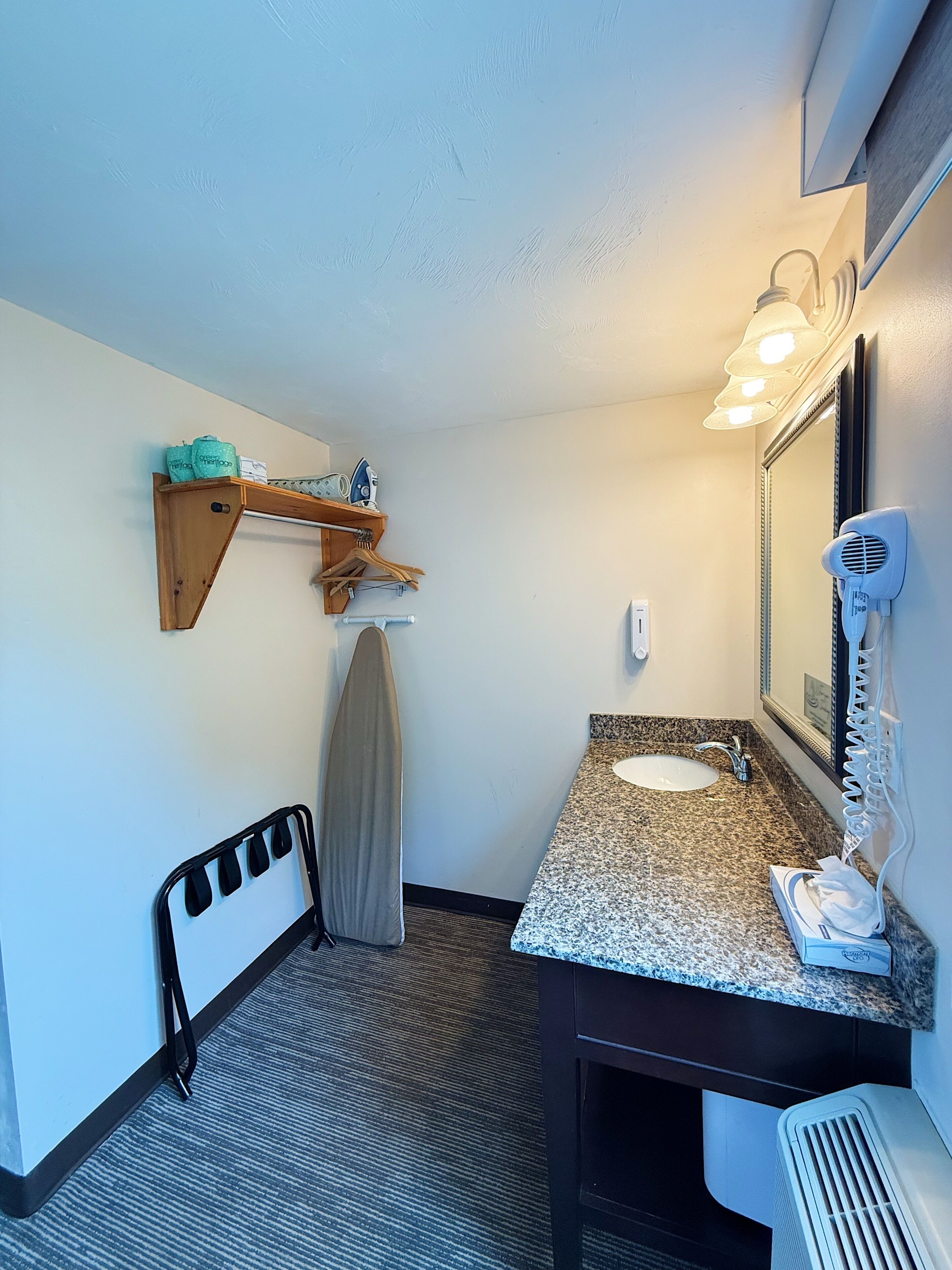 A compact bathroom with a granite countertop sink, a mirror, a hairdryer, and a shelf with personal items.