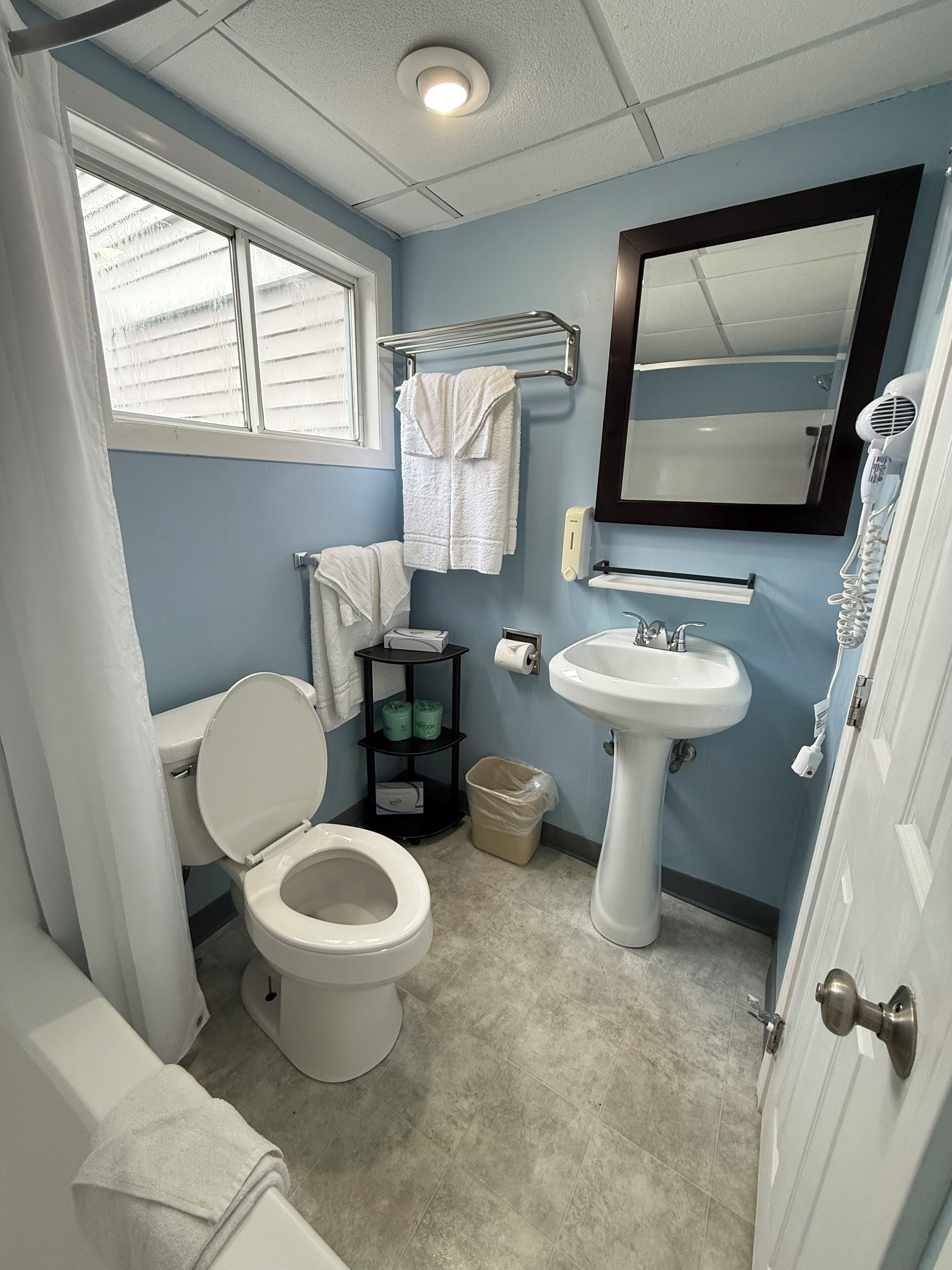 A bathroom with a toilet, sink, and mirror. There is a window with blinds, a towel rack, and a trash can.