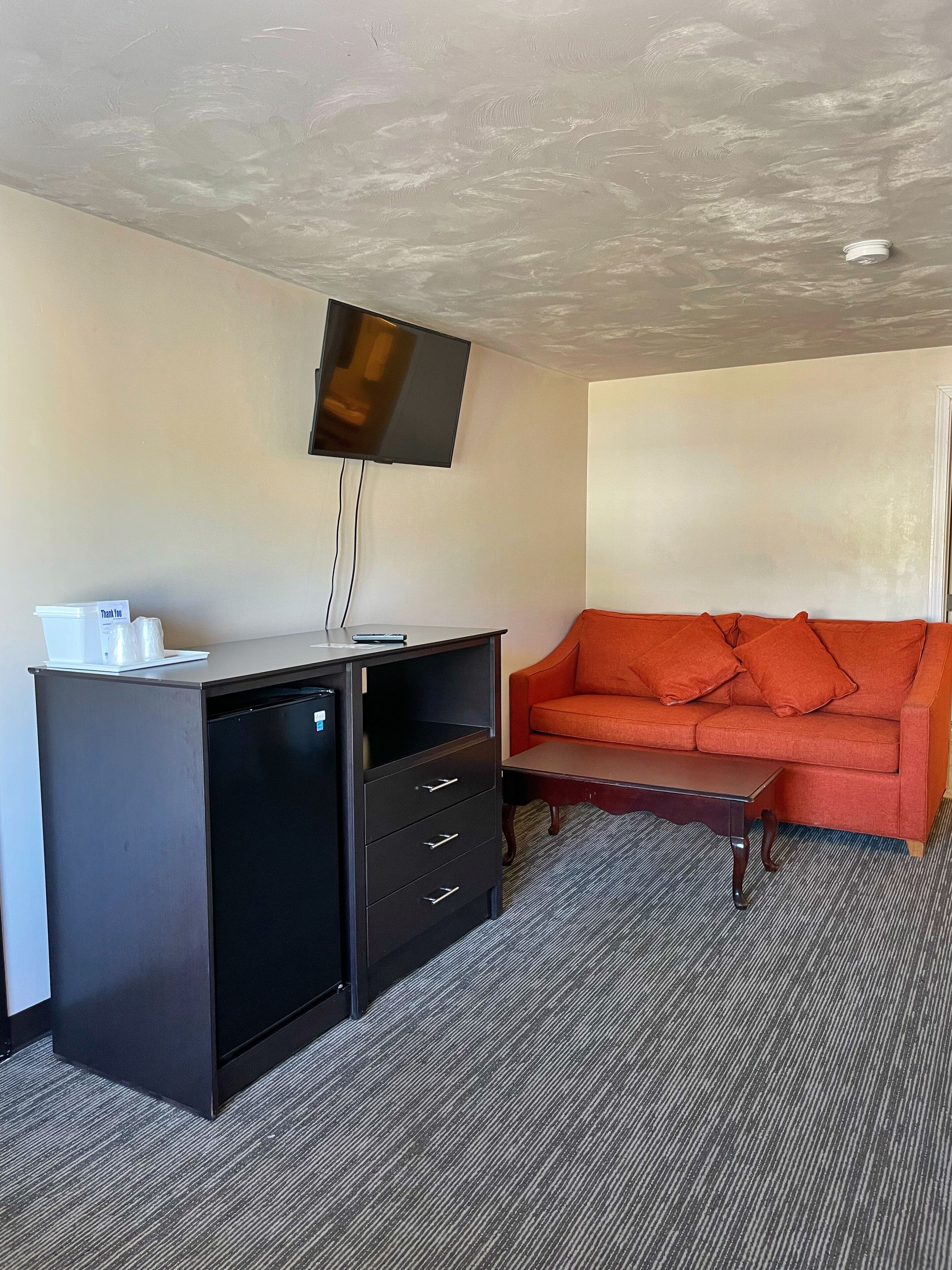 A compact hotel room with a small desk, a television mounted on the wall, a red sofa, and a carpeted floor.