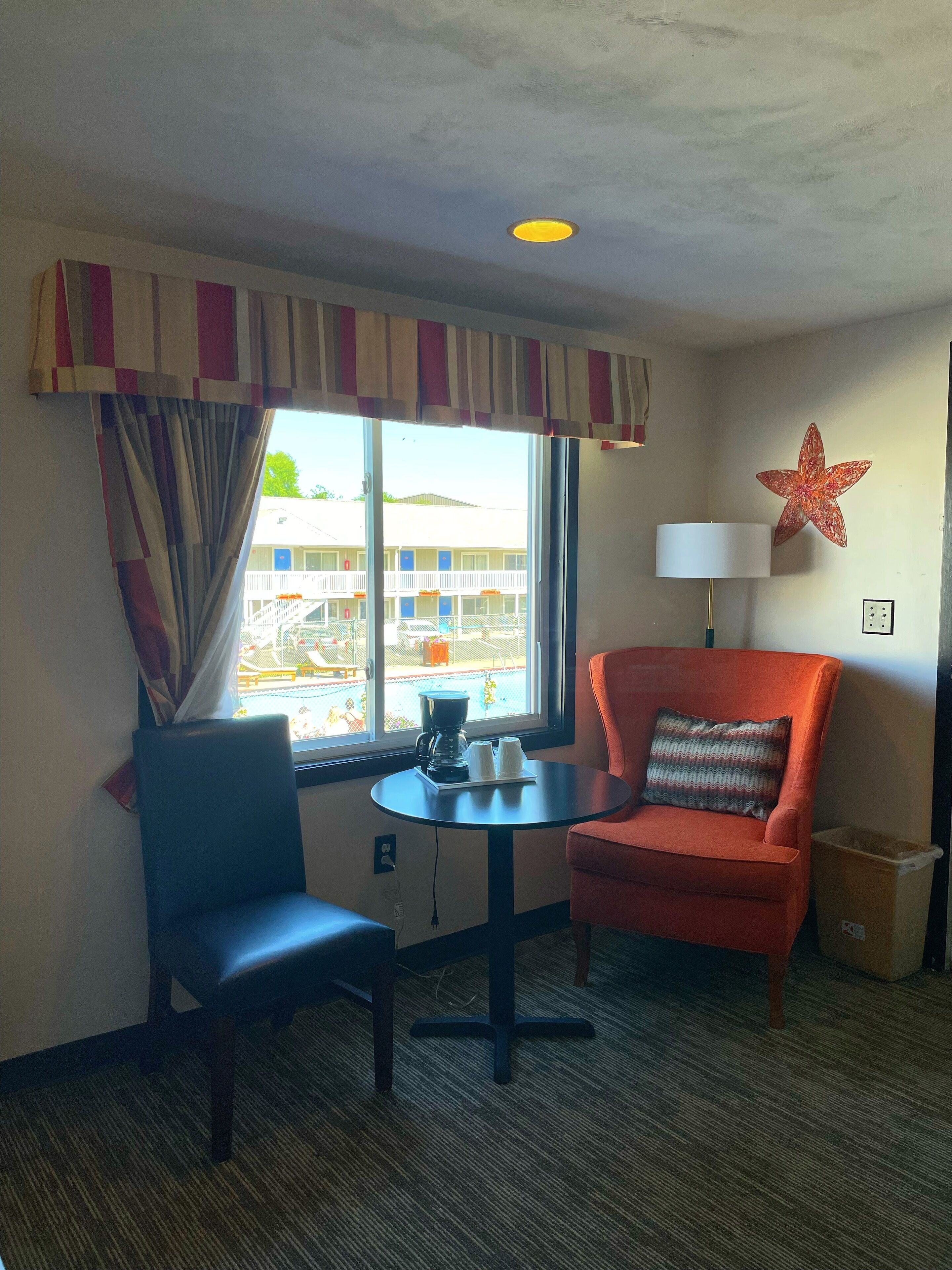 Een hotelkamer met een raam, een tafel met een koffiezetapparaat, twee stoelen, een lamp en een sterren decoratie aan de muur.