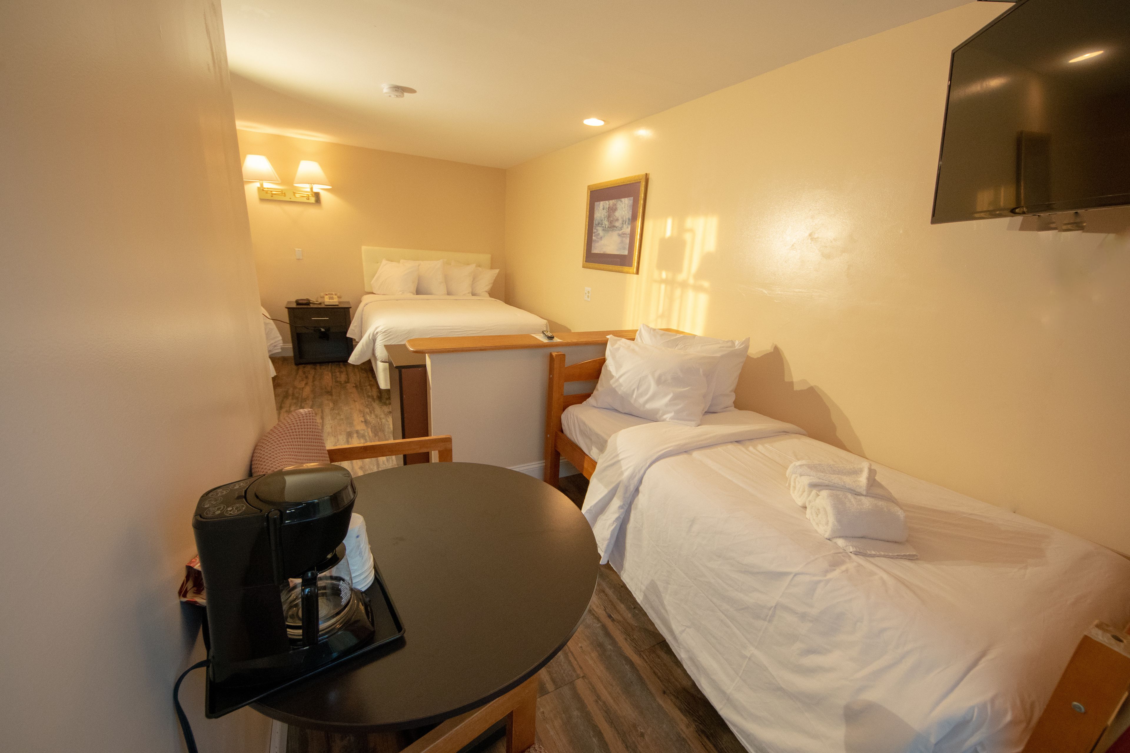 A hotel room with two beds, a small table with a coffee maker, a TV mounted on the wall, and a bedside table with a lamp.